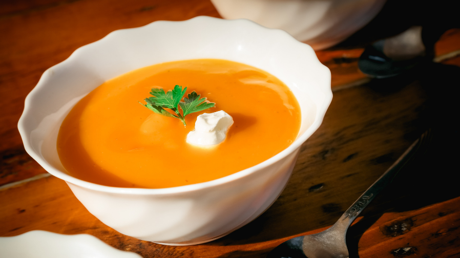 Bowl of smooth orange butternut squash soup topped with a sprig of parsley, with a jar of bone broth and a halved roasted squash behind it