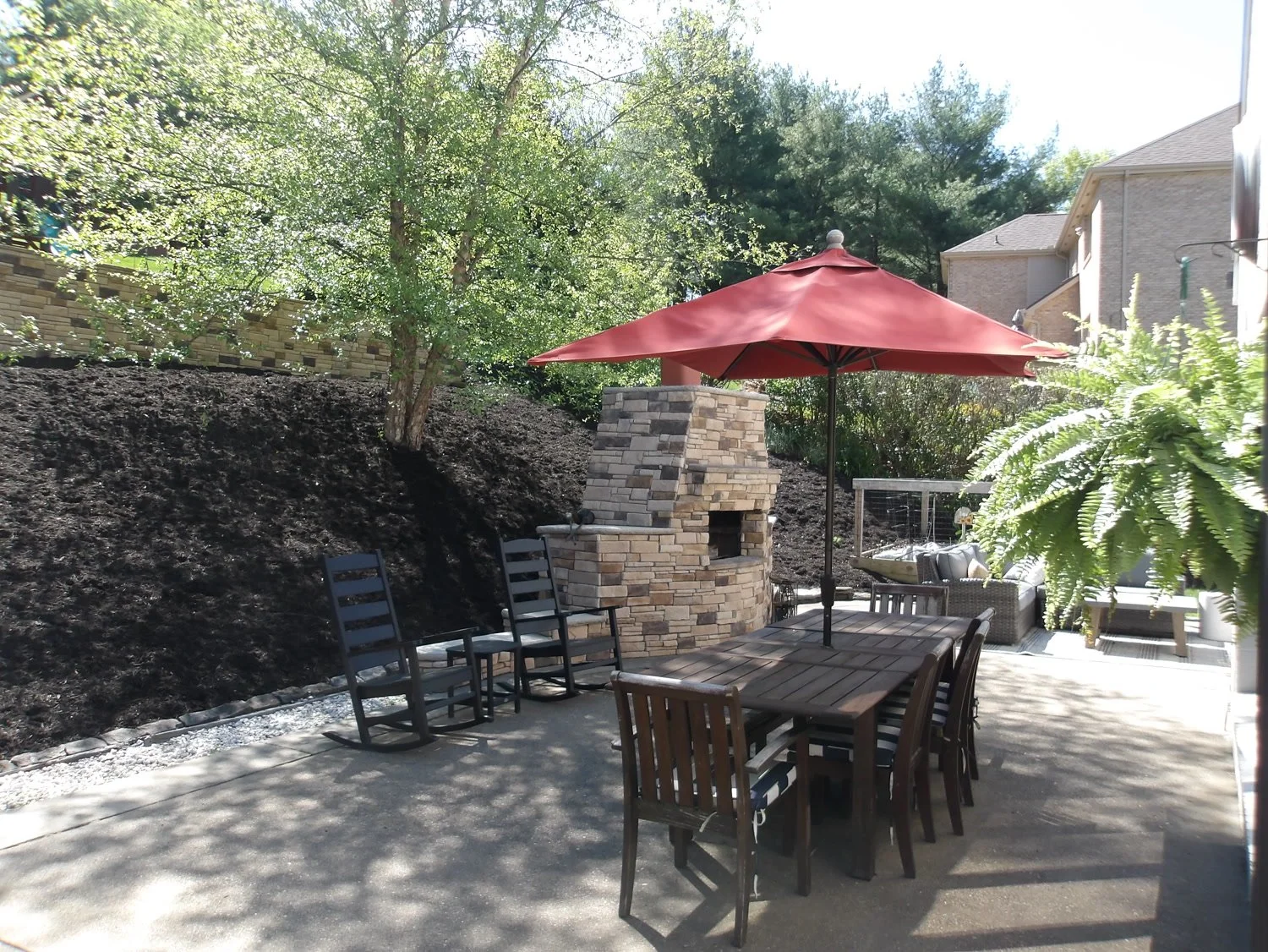 A concrete patio with a dining table, red umbrella and stone fireplace