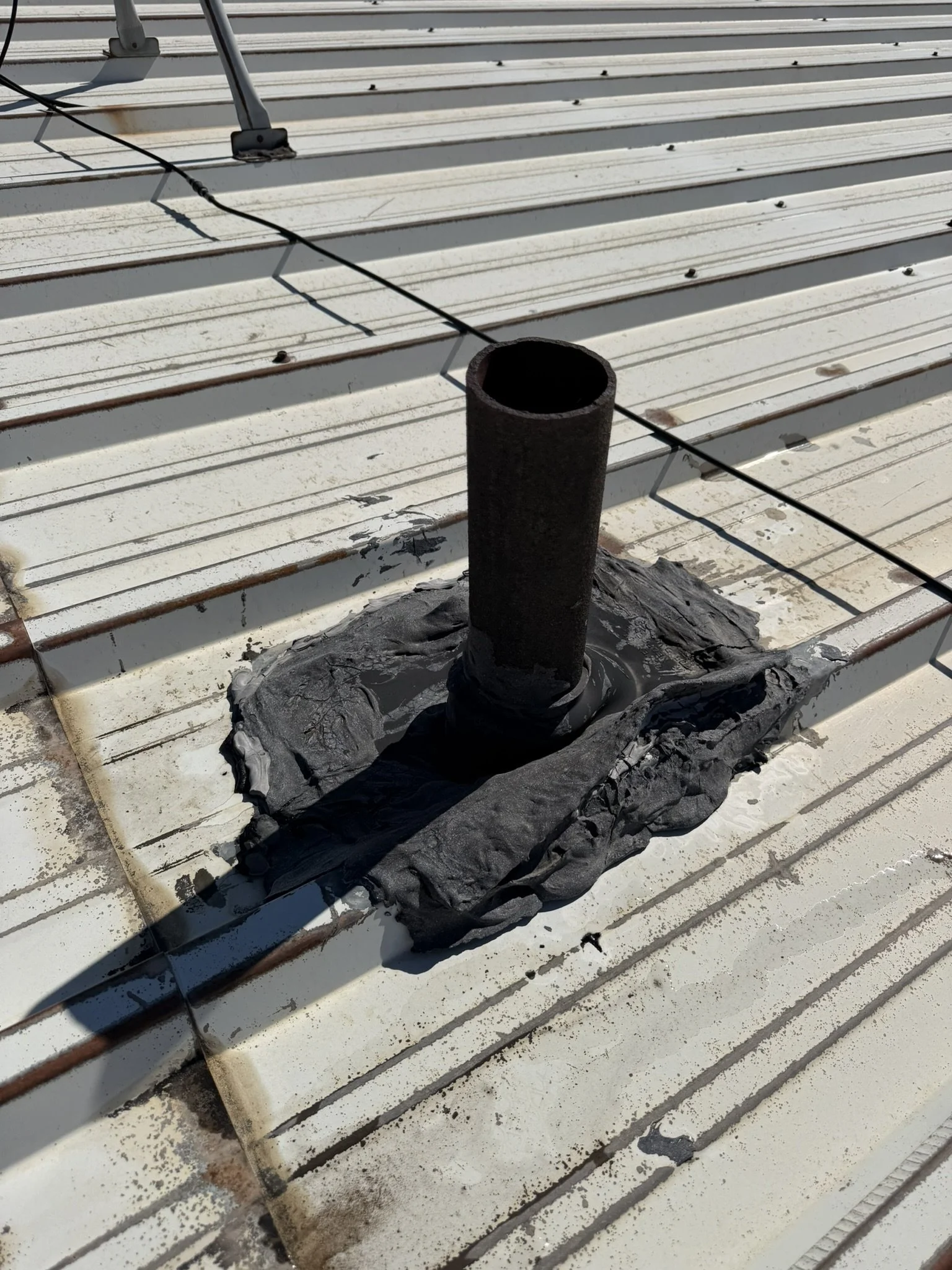 Pipe on a ribbed metal roof surrounded by thick, weathered black sealant.