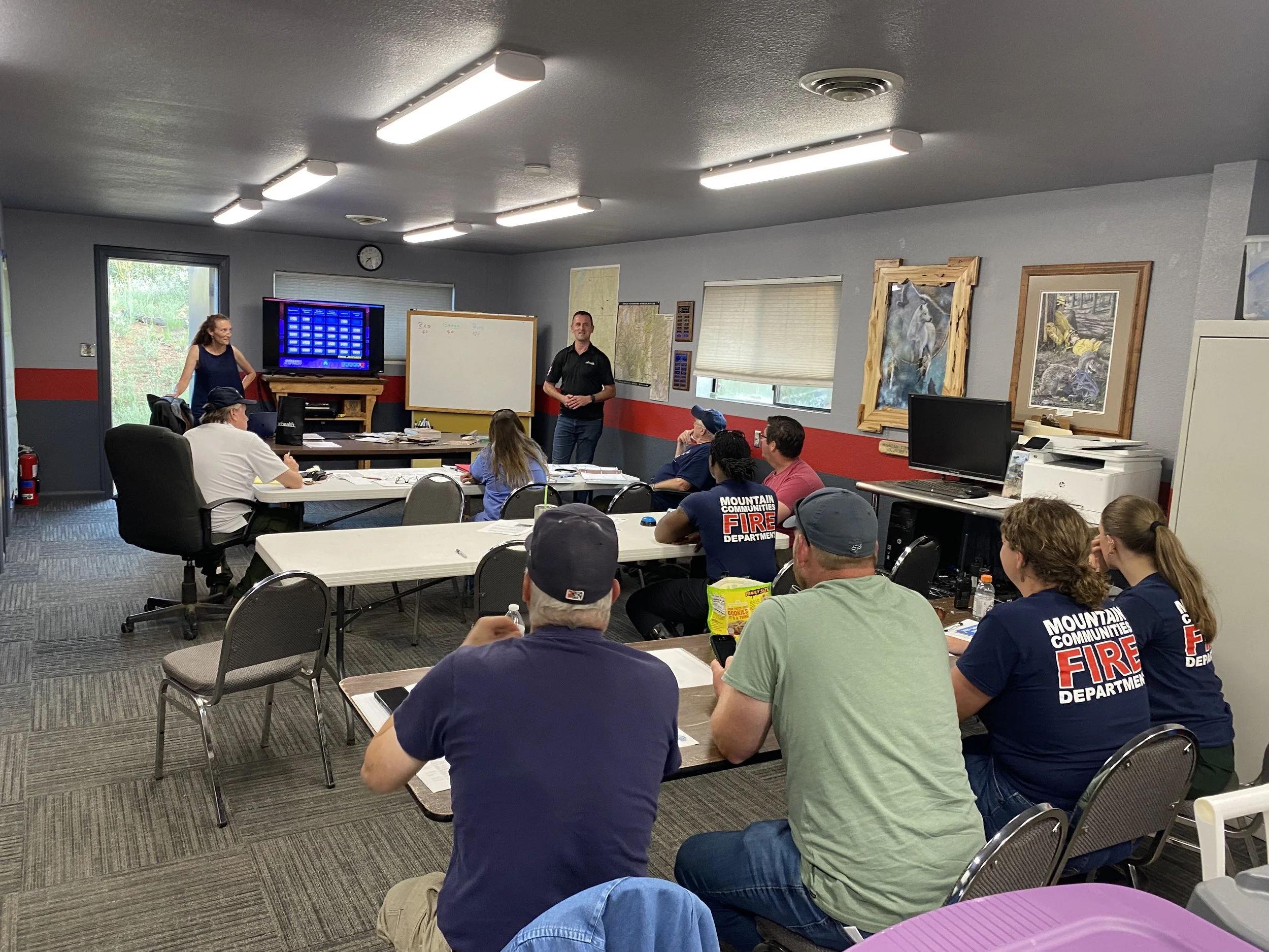 A group of people sitting at tables facing the front of the room while two instructors speak to the group