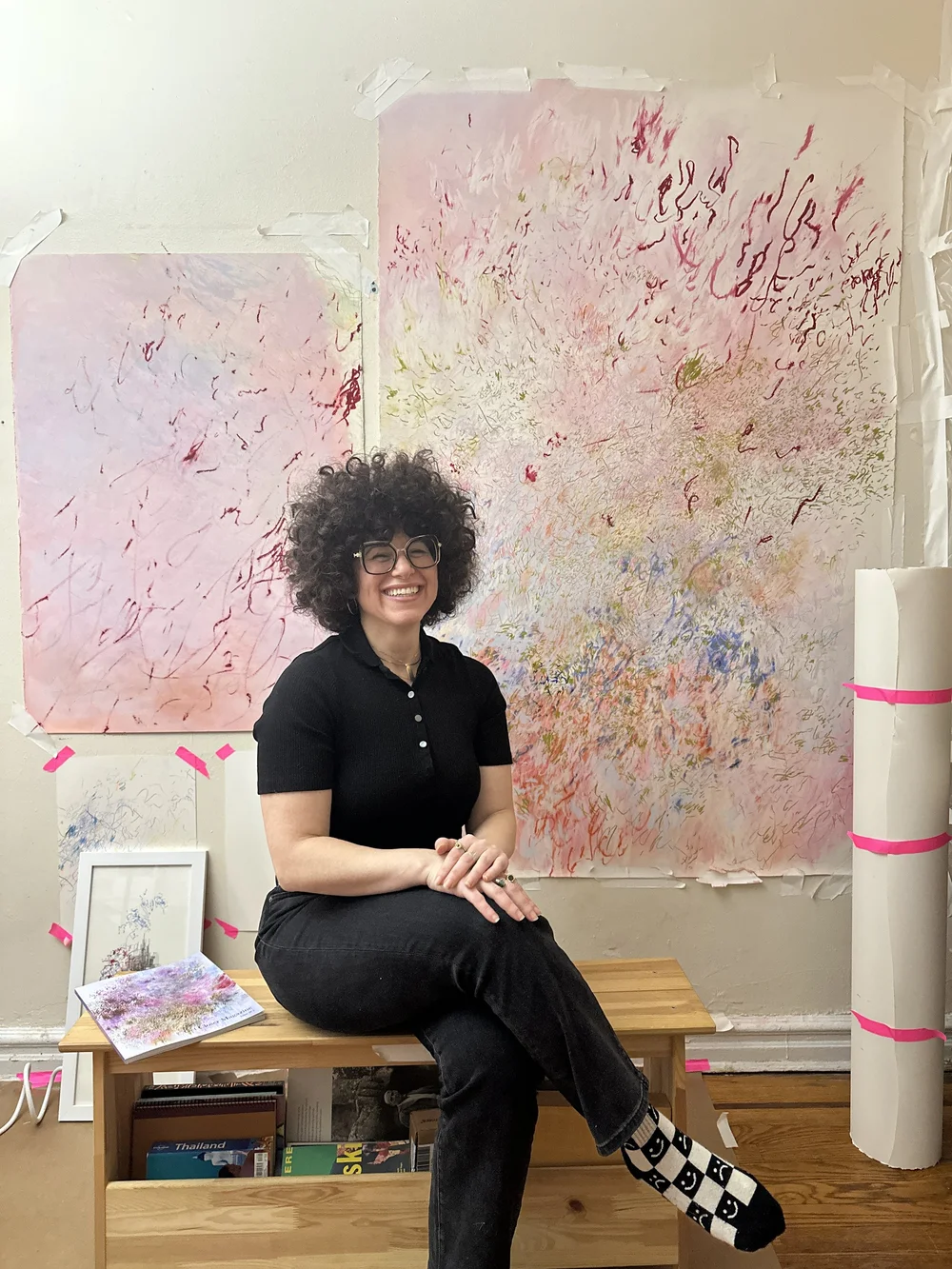 Jalena Hay smiling, seated on a wooden bench in her Brooklyn studio in front of two large pink abstract works on paper.