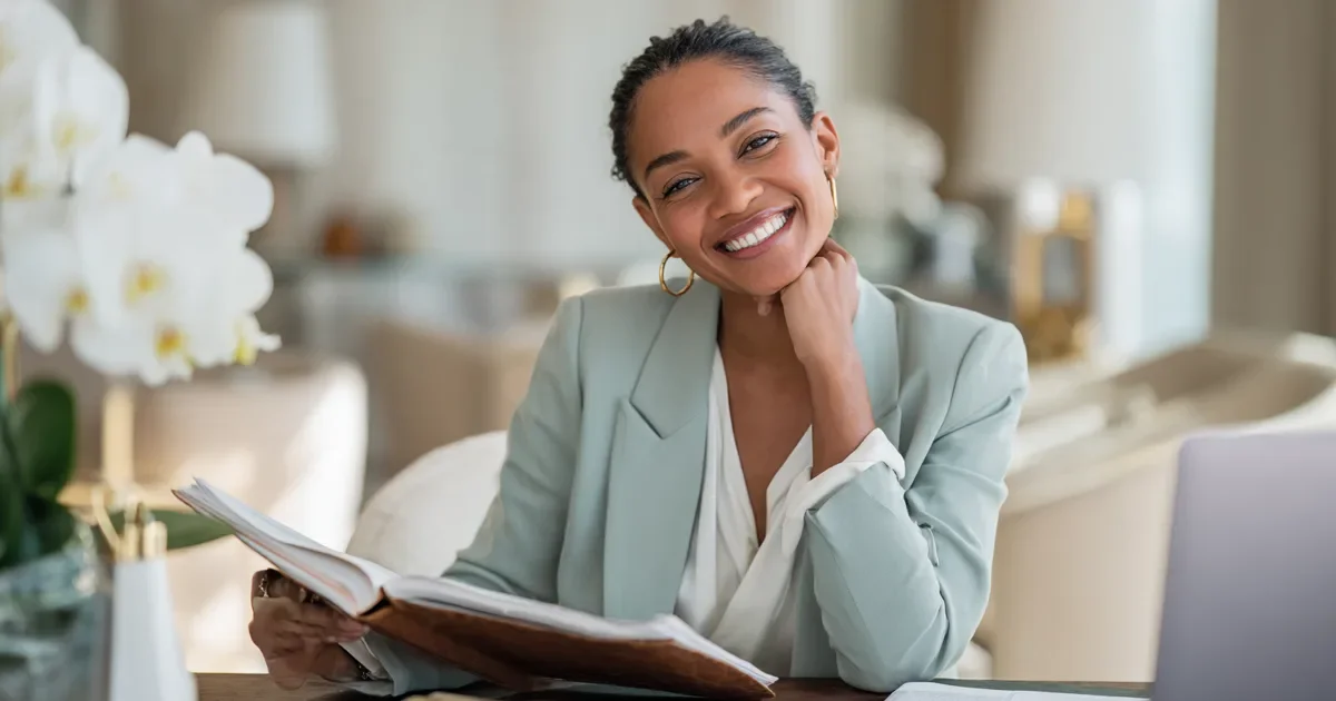 Financial advisor client acquisition cost: a smiling financial advisor in a soft sage blazer reviews a leather planner at a white oak desk in a bright, airy office with white orchids and cream linen chairs.