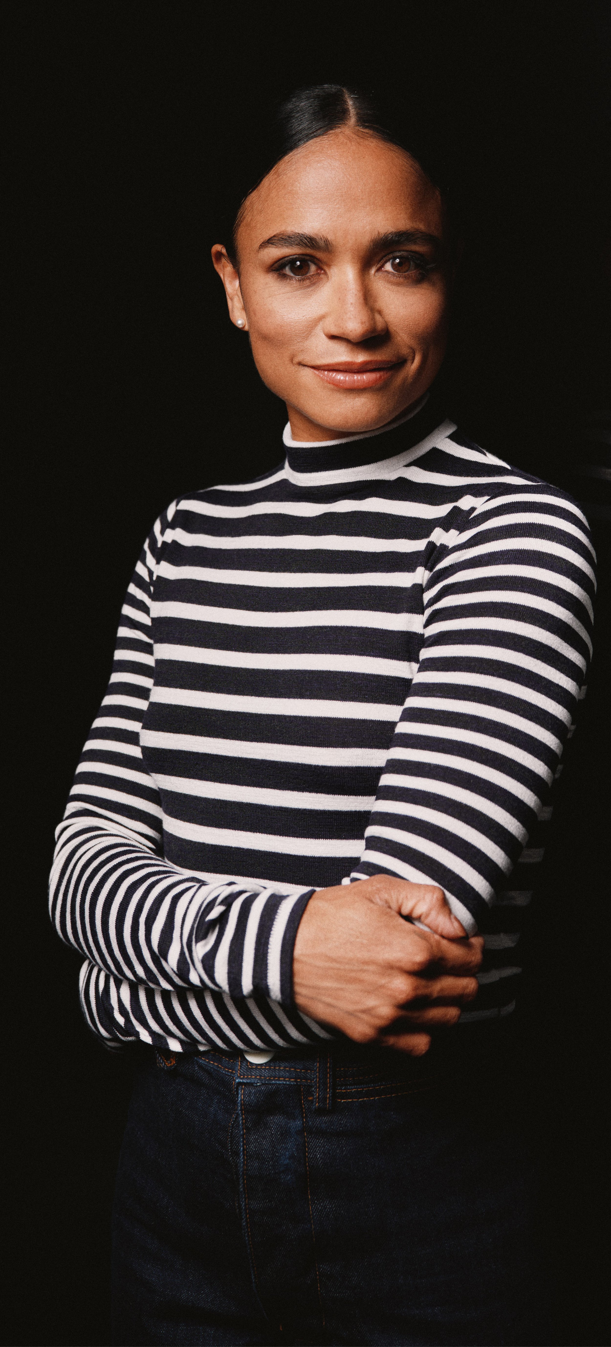 A light skinned Black and Mexican American woman with slicked back black hair, wearing a black and white striped low turtleneck sweater and dark blue jeans, smiling.