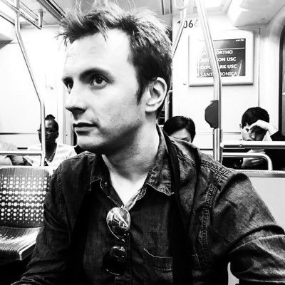 A black and white headshot of a white man with short dark hair sitting on public transportation, wearing a denim shirt with aviator sunglasses hanging from his collar