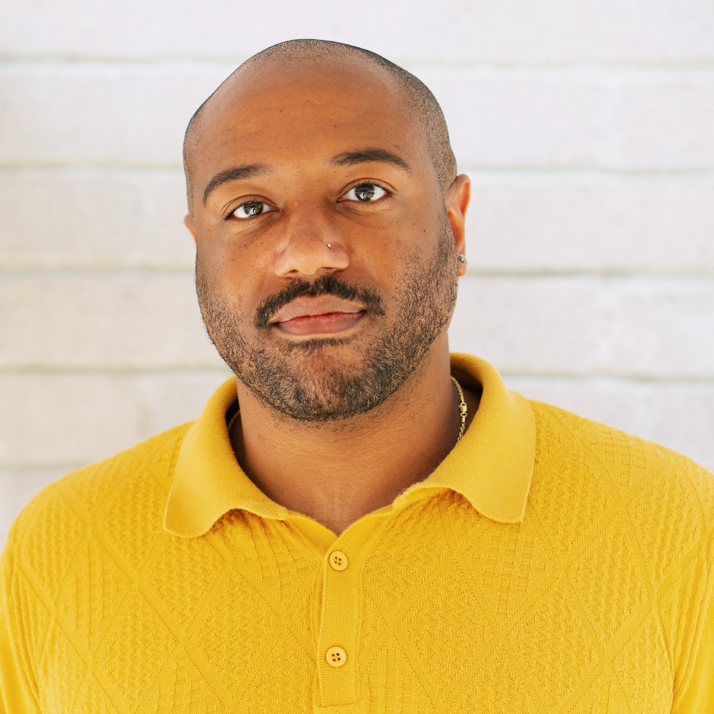 A Black bald and a short black beard wearing a yellow knit collared shirt.