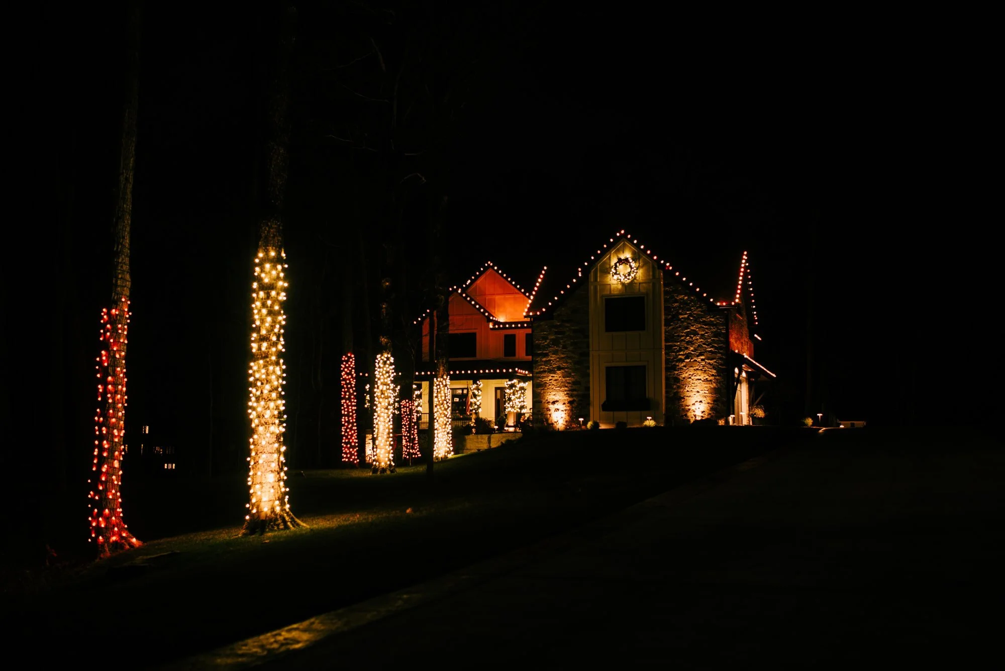 Tall tree trunks wrapped in warm white and red holiday lights beside a home