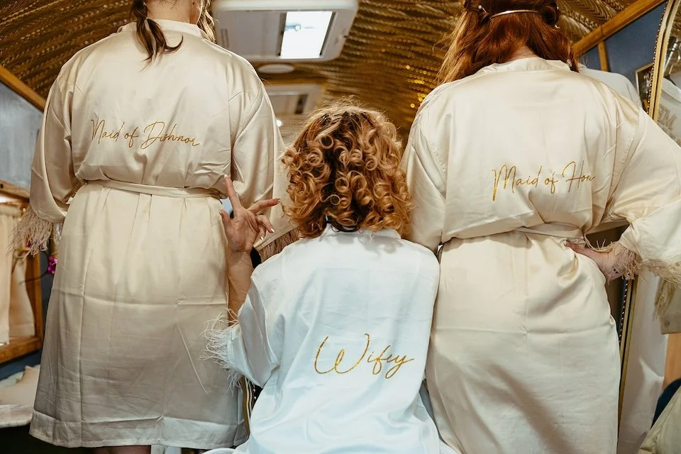 Three custom satin robes reading 'Wifey,' 'Maid of Dishnor,' and 'Maid of Honor' inside the Stargazer Social Club suite.