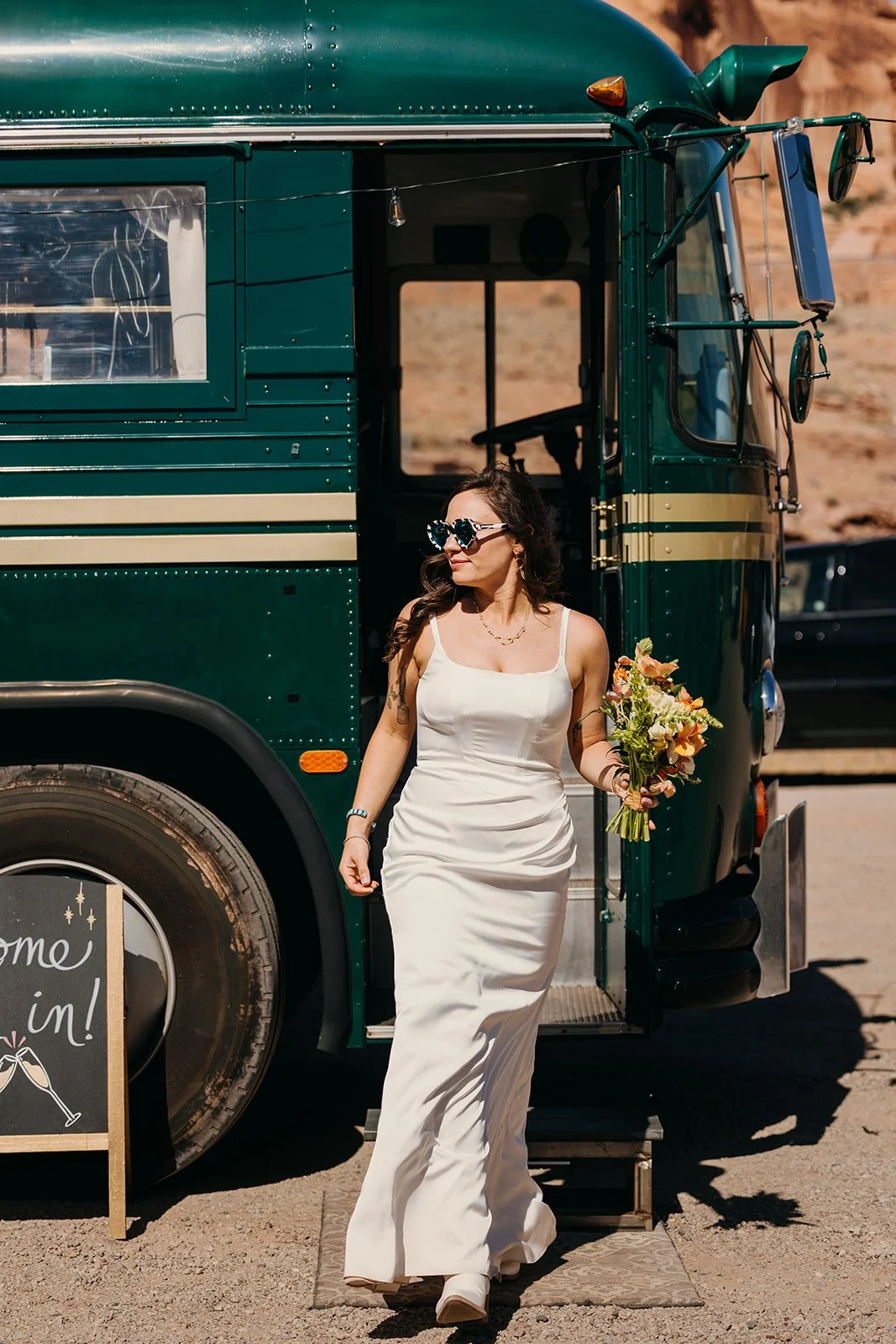 A bride in a white gown and sunglasses stepping off the Stargazer bus, holding a wildflower bouquet with red-rock cliffs behind her.