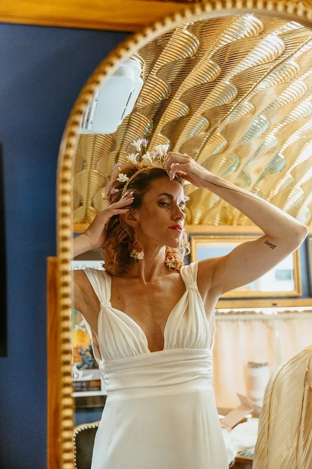 A bride in a white gown adjusting her floral crown in front of a gilded arch mirror inside the Stargazer Social Club suite.