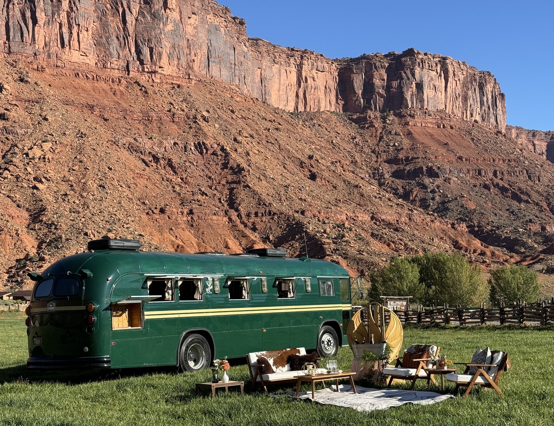 Regina, Stargazer's vintage green Crown bus, parked on the grass beneath the red cliffs with a curated lounge setup in the foreground