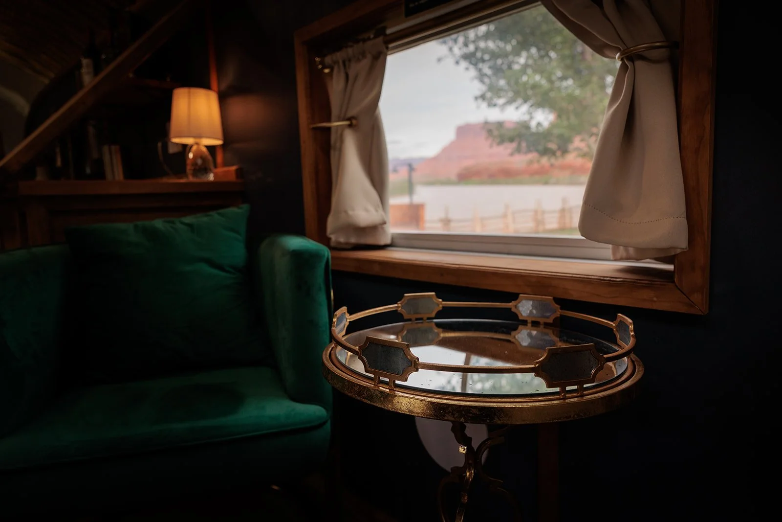 Inside Regina: a teal velvet sofa, a brass-rimmed mirrored side table, and a wood-framed window opening onto the Colorado River and red cliffs beyond