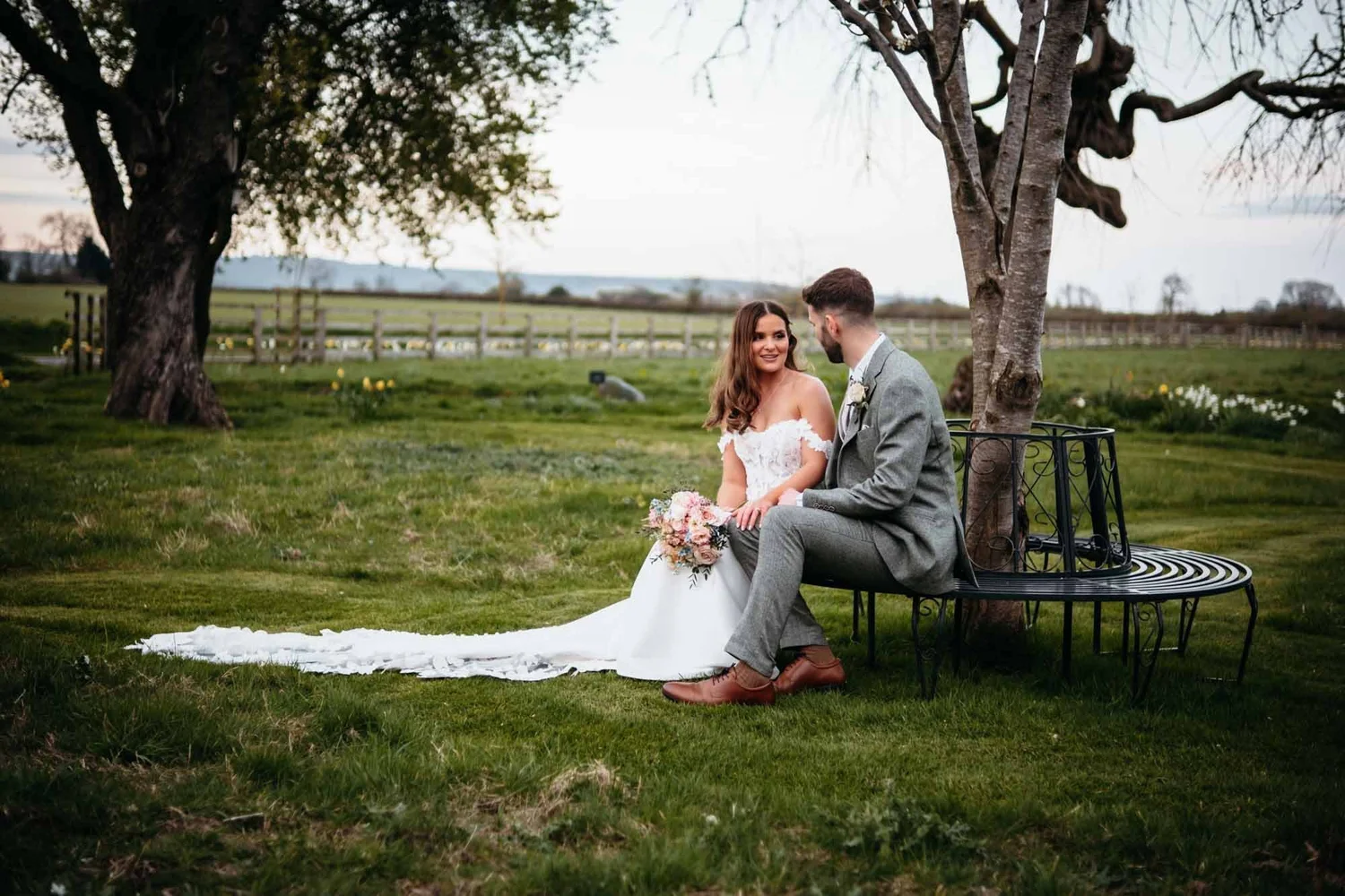 Couple on a bench beneath the trees with the wedding dress spread out, Thirsk Lodge Barns
