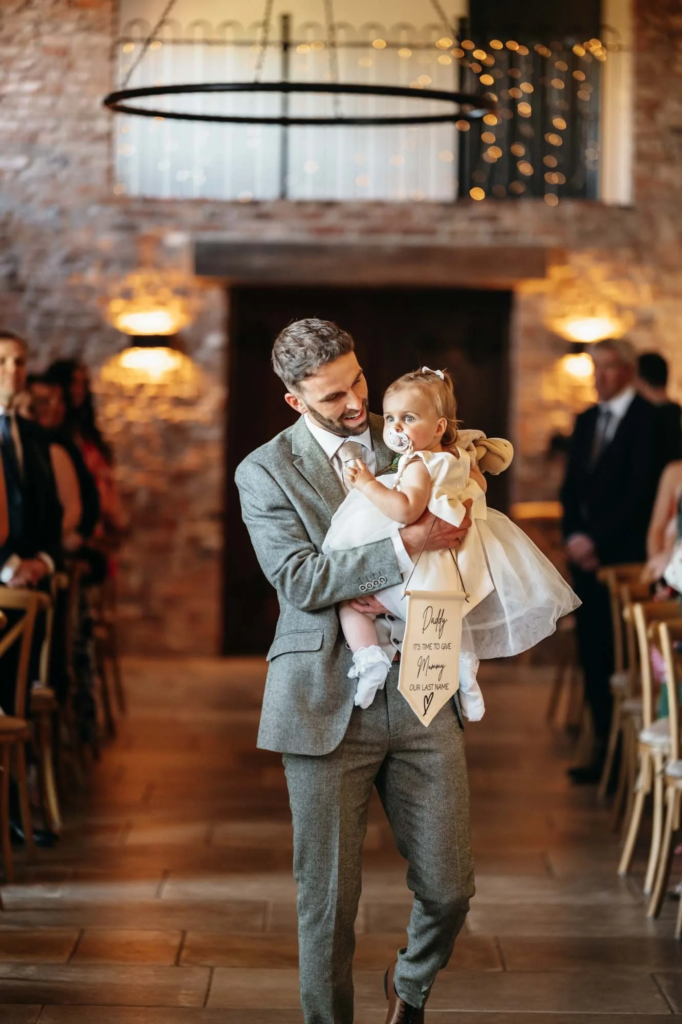 Groom carrying a small child in a white dress down the aisle of the Main Barn at Thirsk Lodge Barns