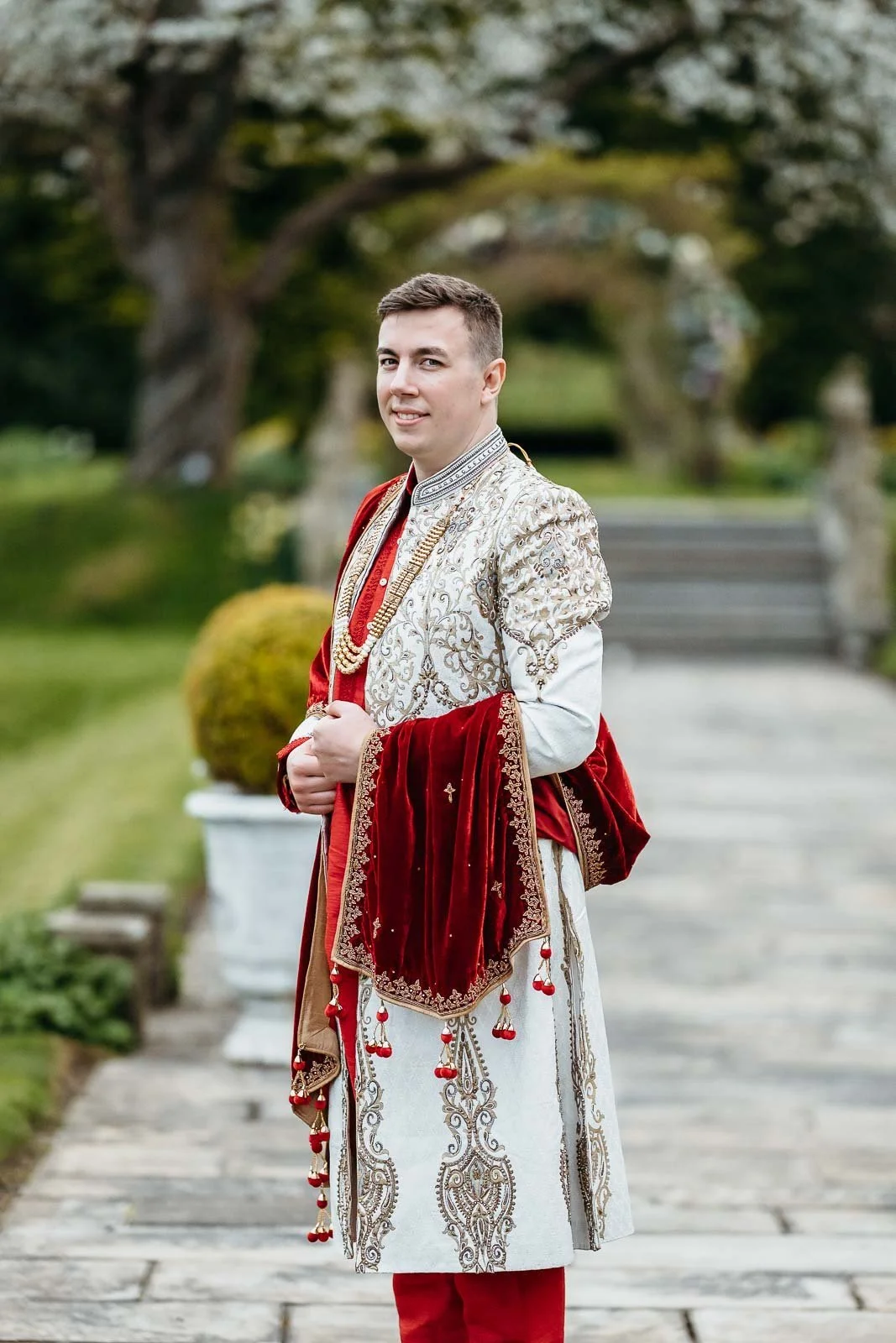 Groom in a cream and red sherwani on a stone garden path at Lartington Hall