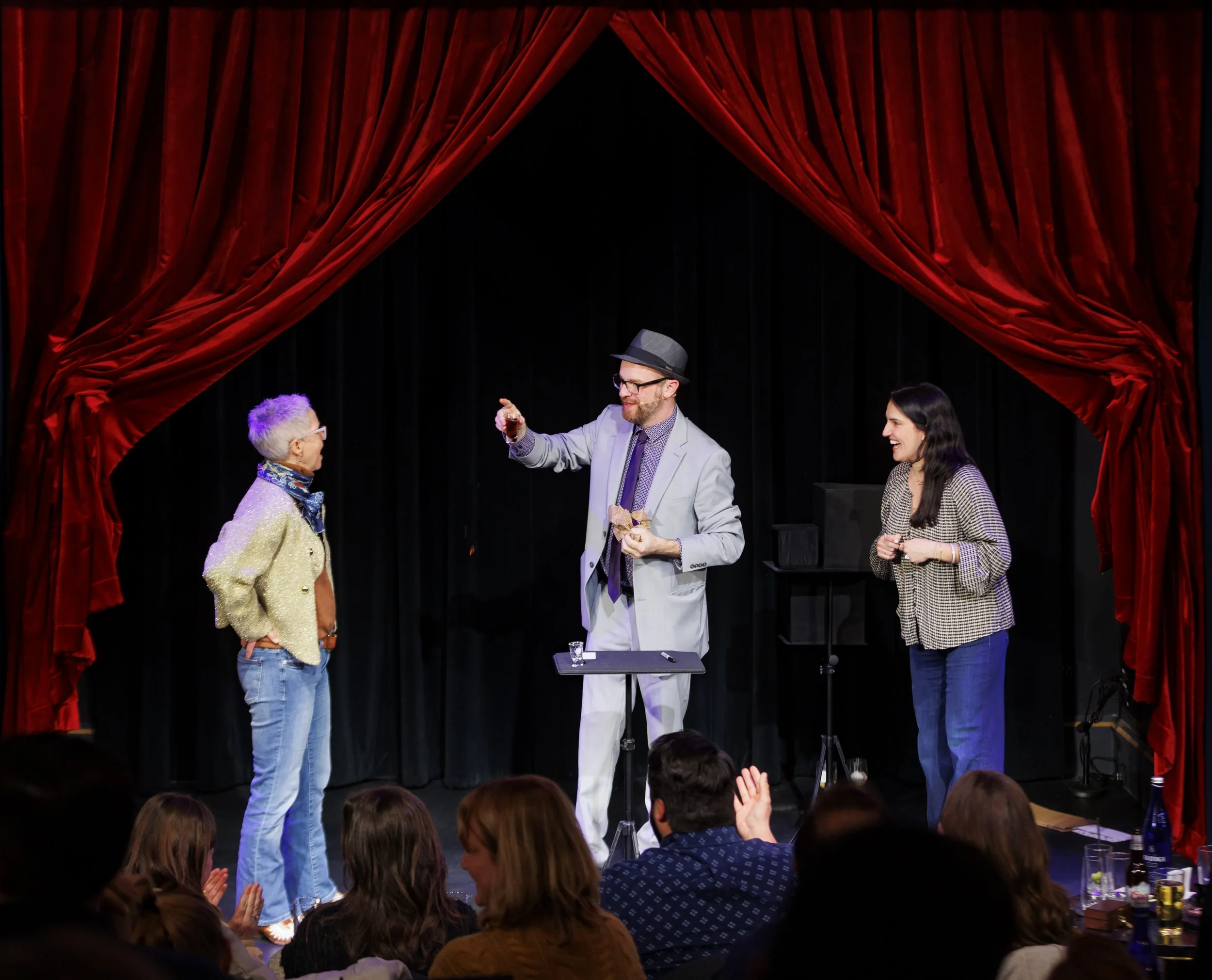 Magician performing on stage with two guests in front of a red curtain