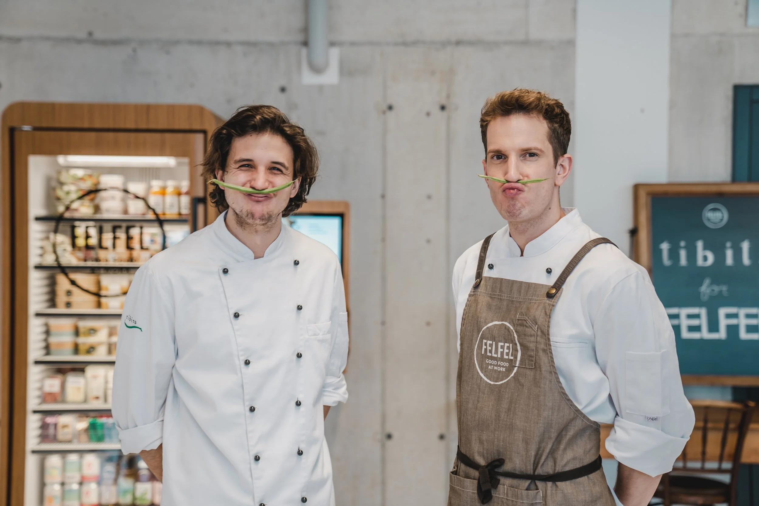 Zwei Partnerköche von FELFEL mit Bohnen-Schnurrbart vor dem FELFEL Kühlschrank