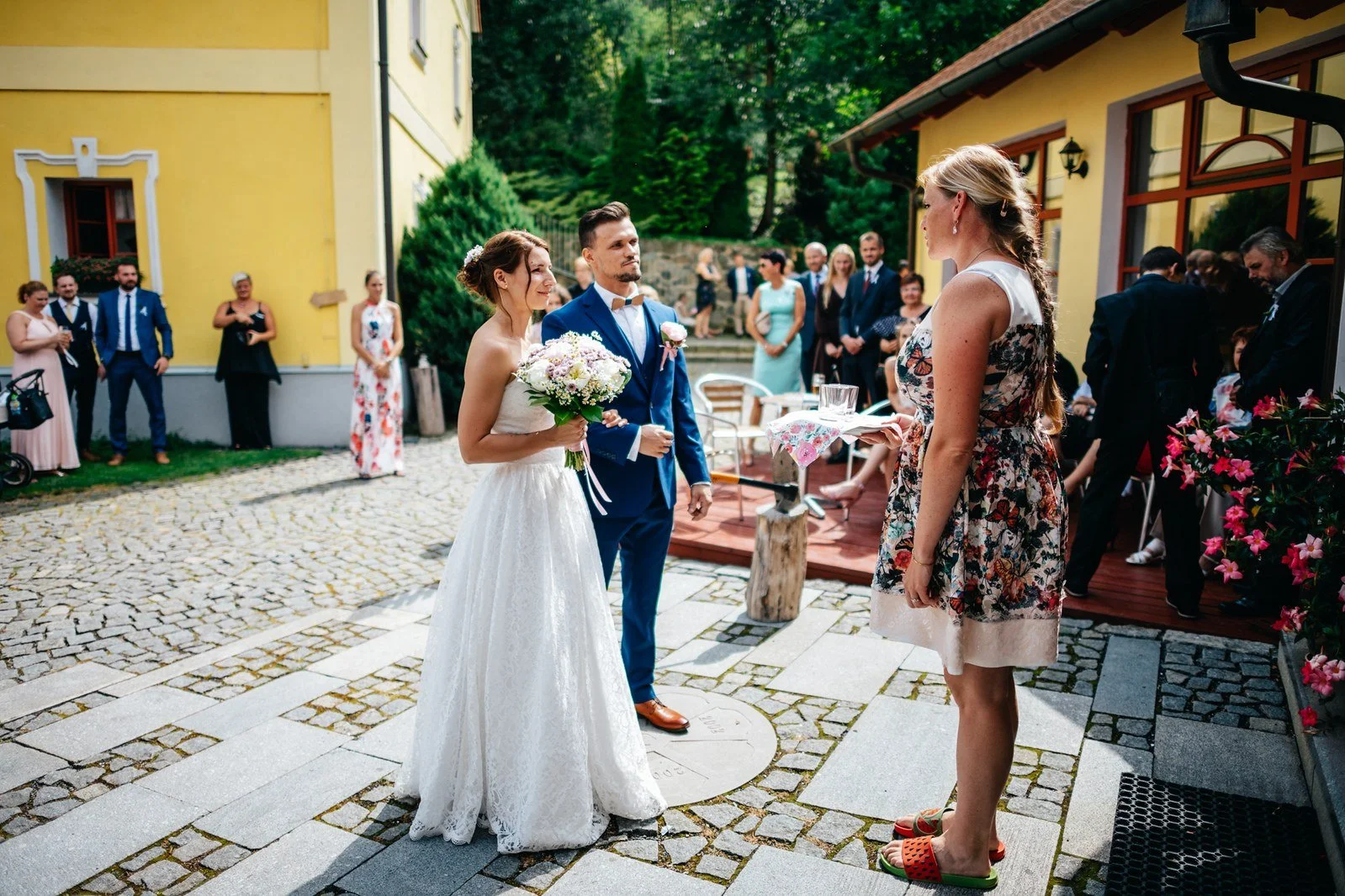 Postava ve svatebních šatech vítá hosta u venkovního posezení.