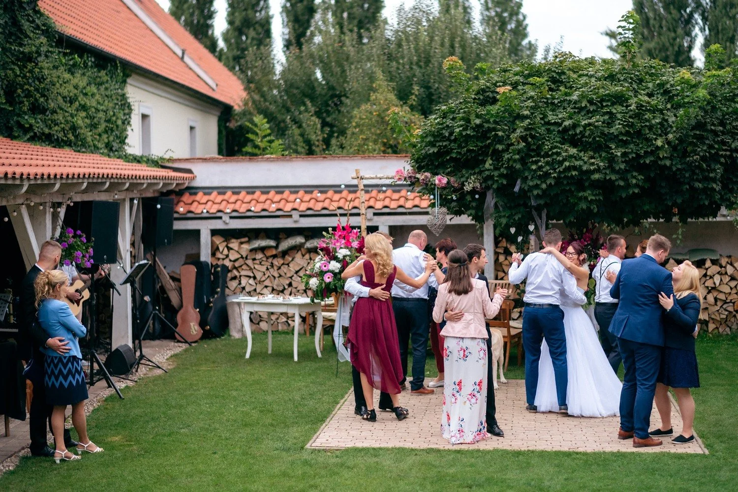 Několik dvojic tančí na dvoře mezi kamennými zdmi a stromy.