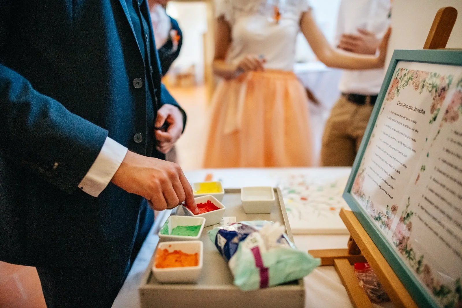 Detail ženichovy ruky u barevných polštářků a zarámované tabulky na stole.