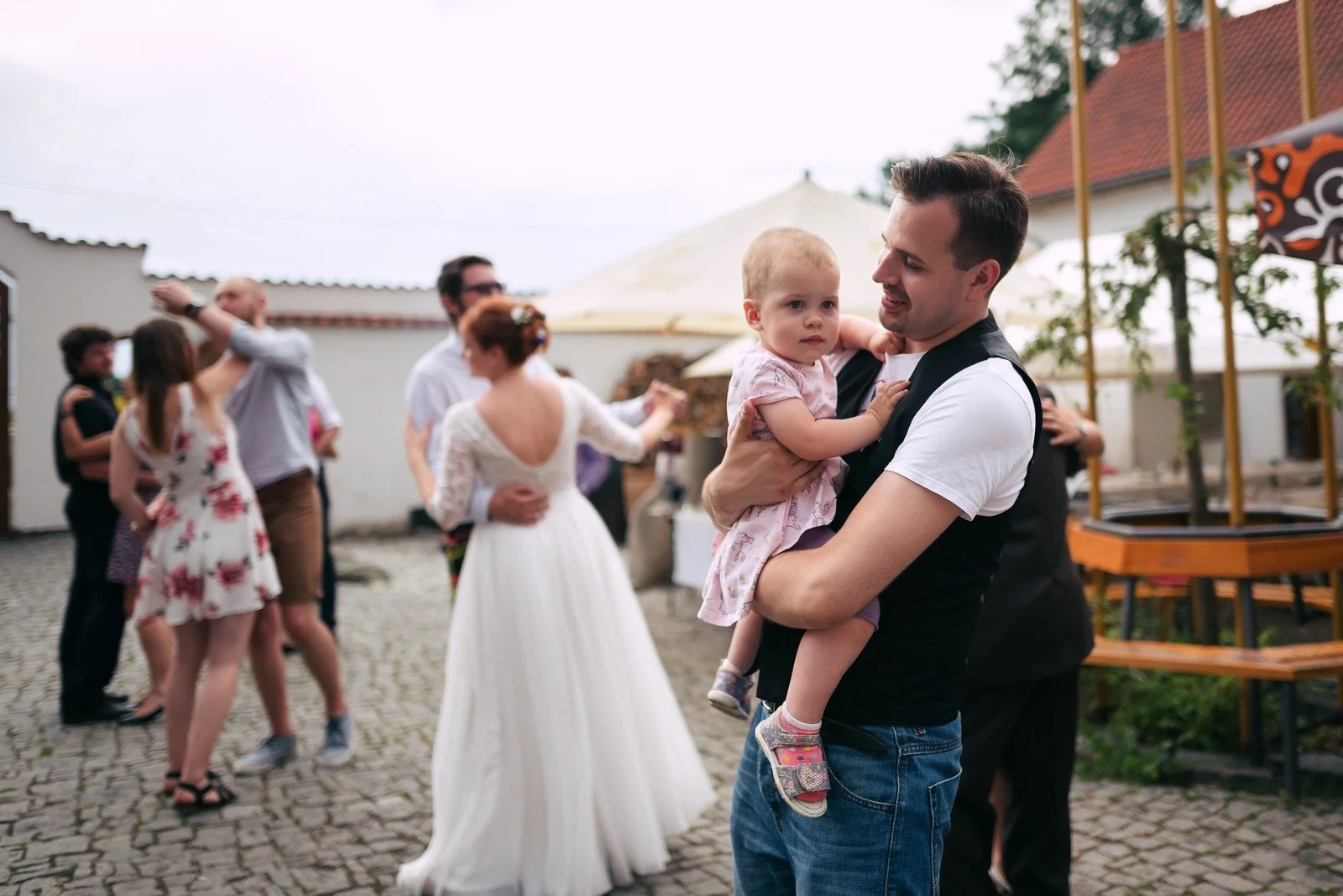 Muž drží v náručí malé dítě, zatímco vedle něj stojí žena v bílých šatech.