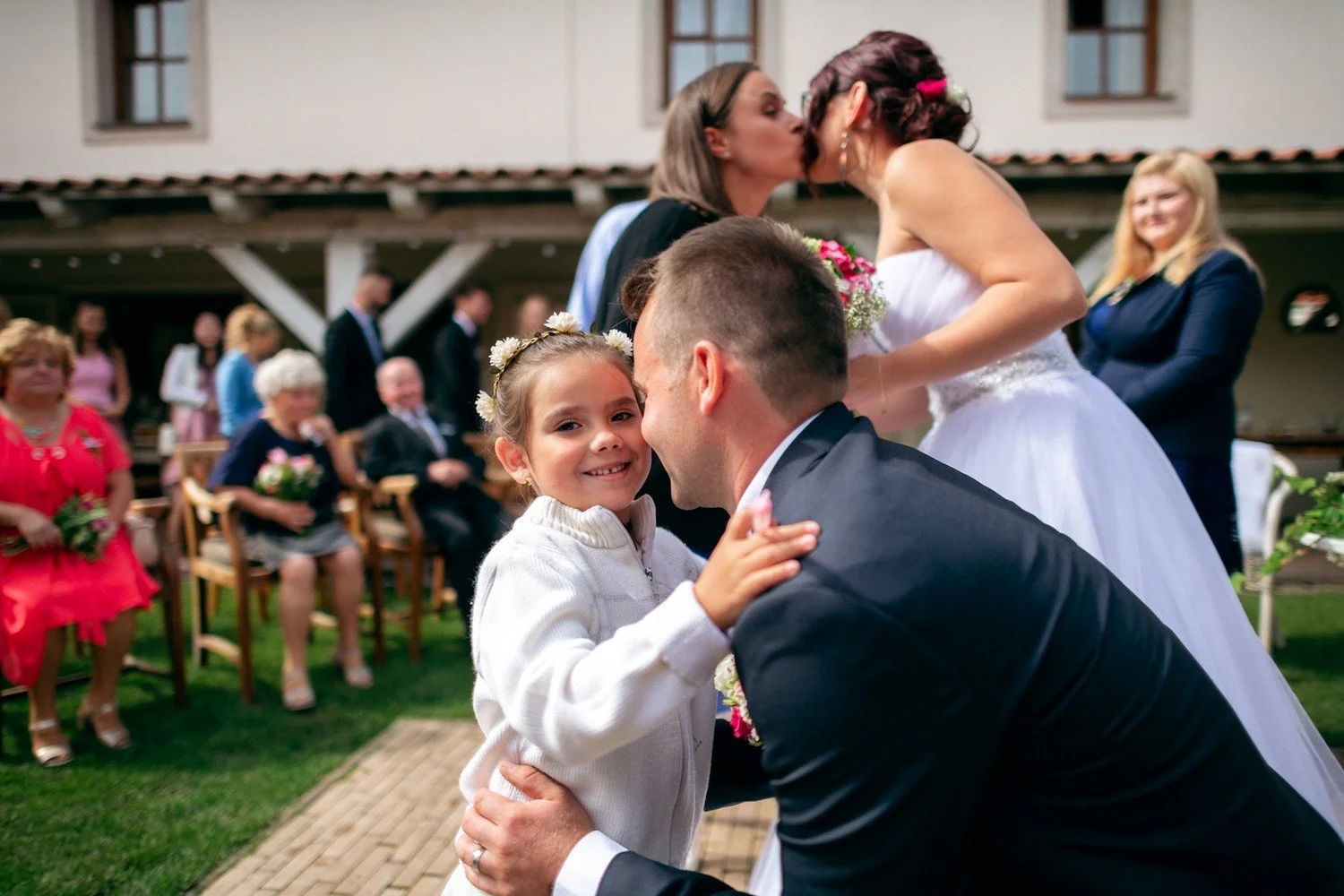 Usměvavá dívka objímá muže v obleku, zatímco se v pozadí zdraví dvě ženy.
