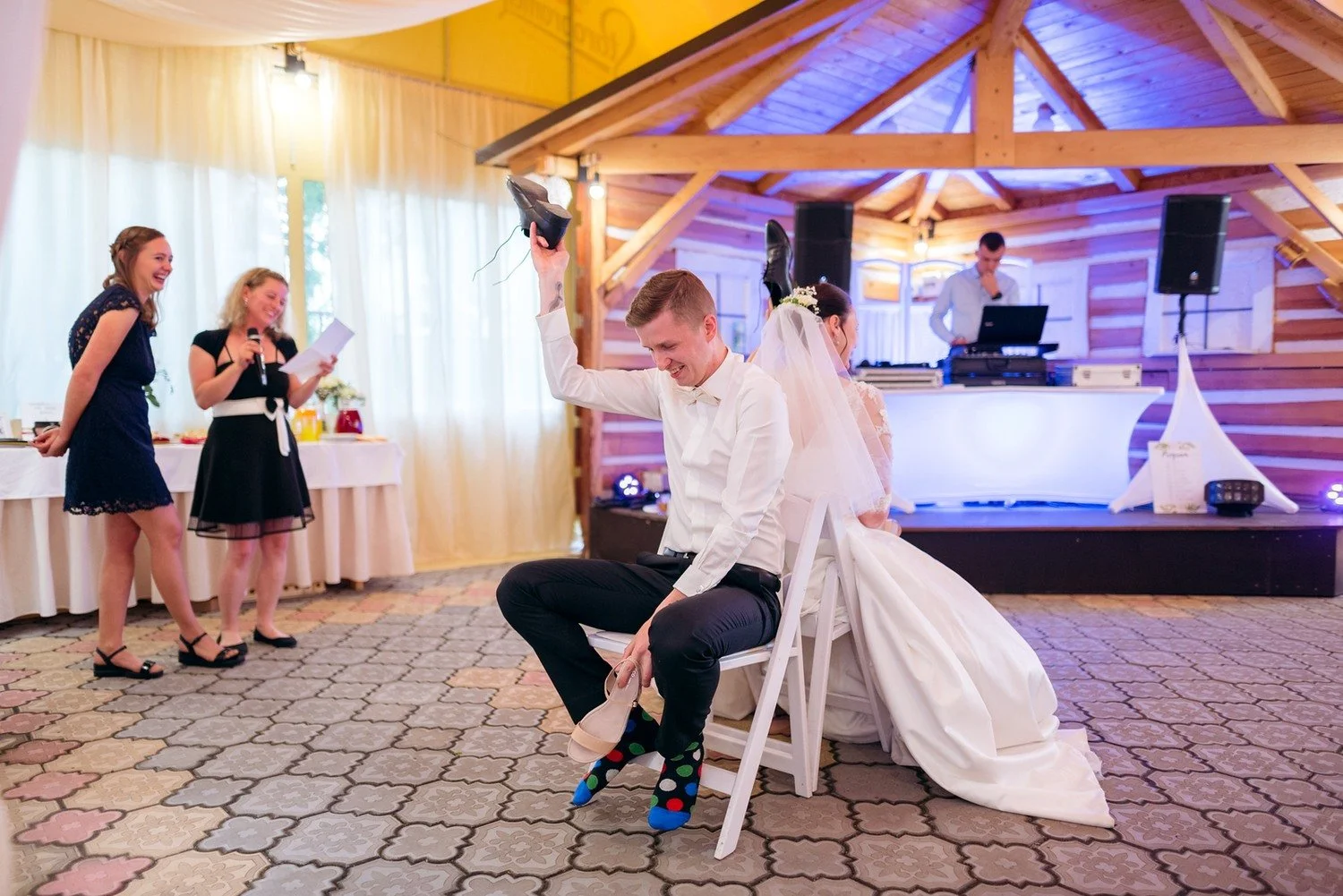 Muž se sklání u lemu svatebních šatů a odhaluje barevné ponožky, okolní ženy scénu se smíchem sledují.