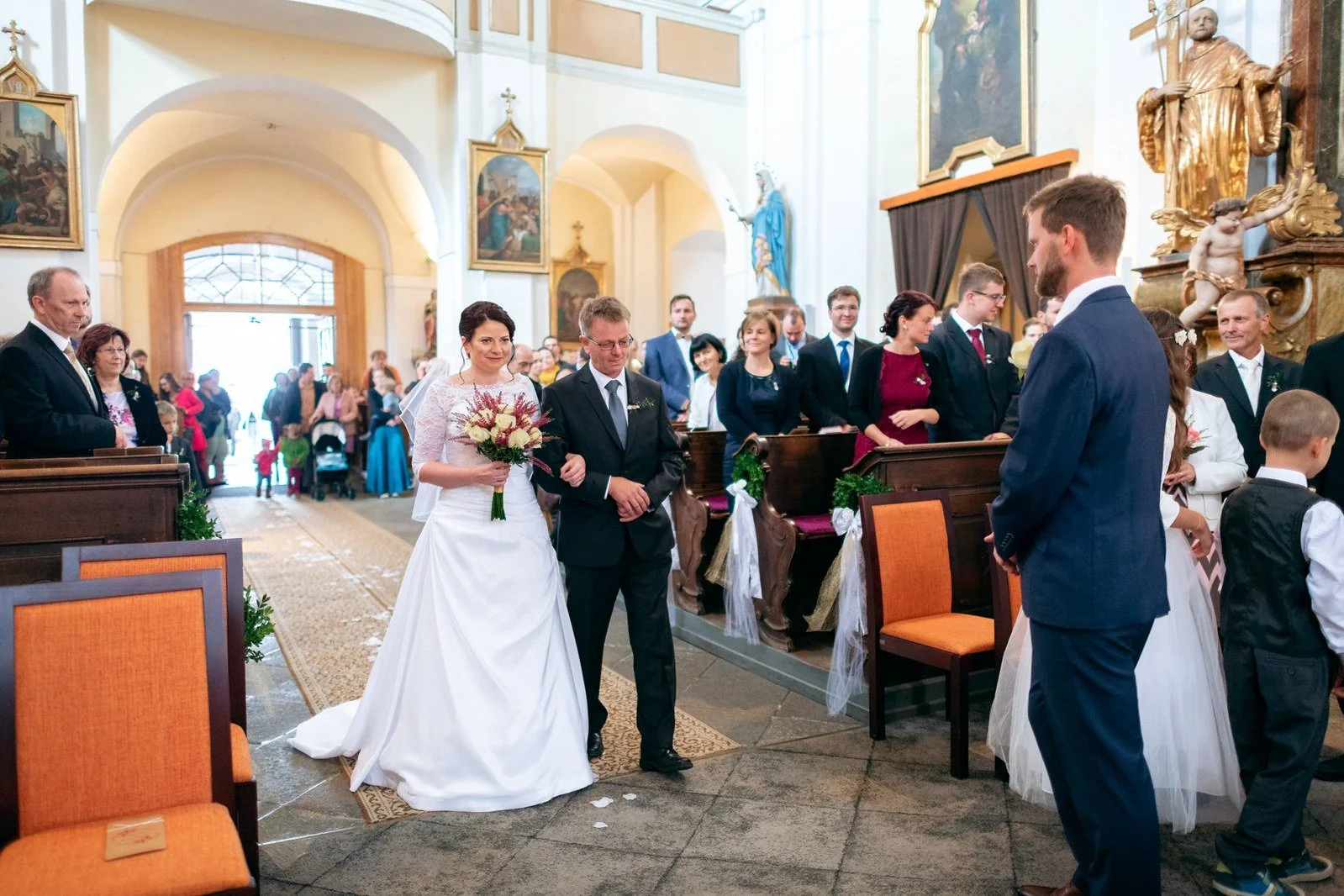 Žena v dlouhých bílých šatech kráčí uličkou kostela po boku muže v tmavém obleku.