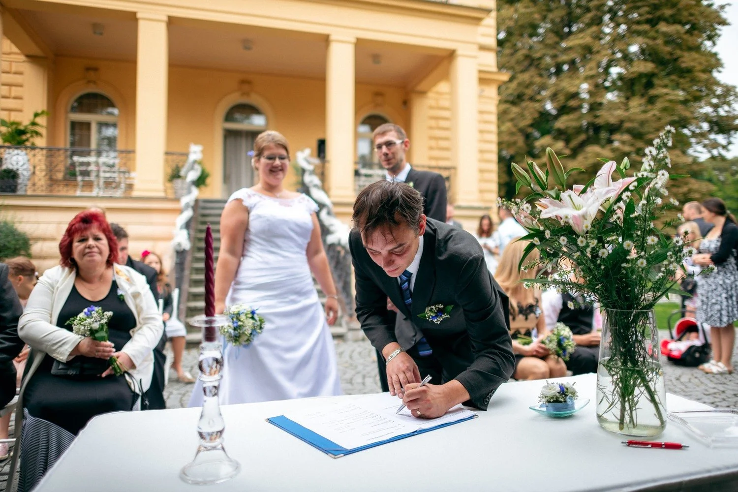 Nevěsta stojí u stolu, zatímco ženich dokončuje podpis.
