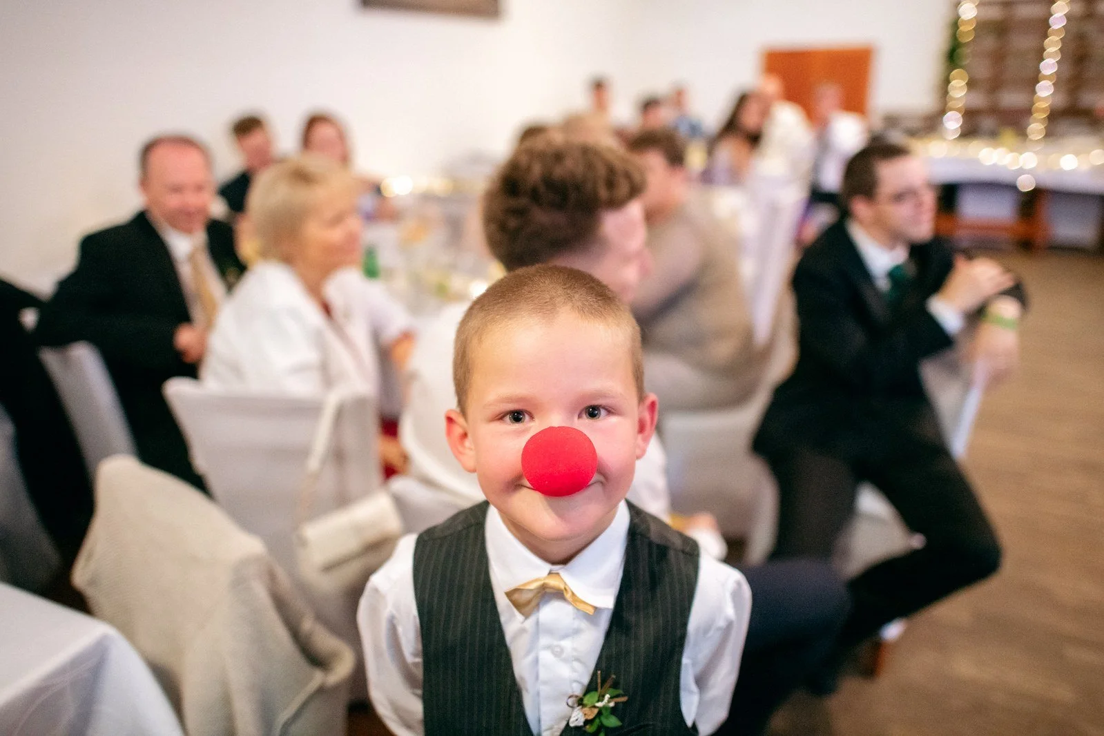 Chlapec ve vestě a motýlku hledí přímo do objektivu s červeným klaunským nosem.