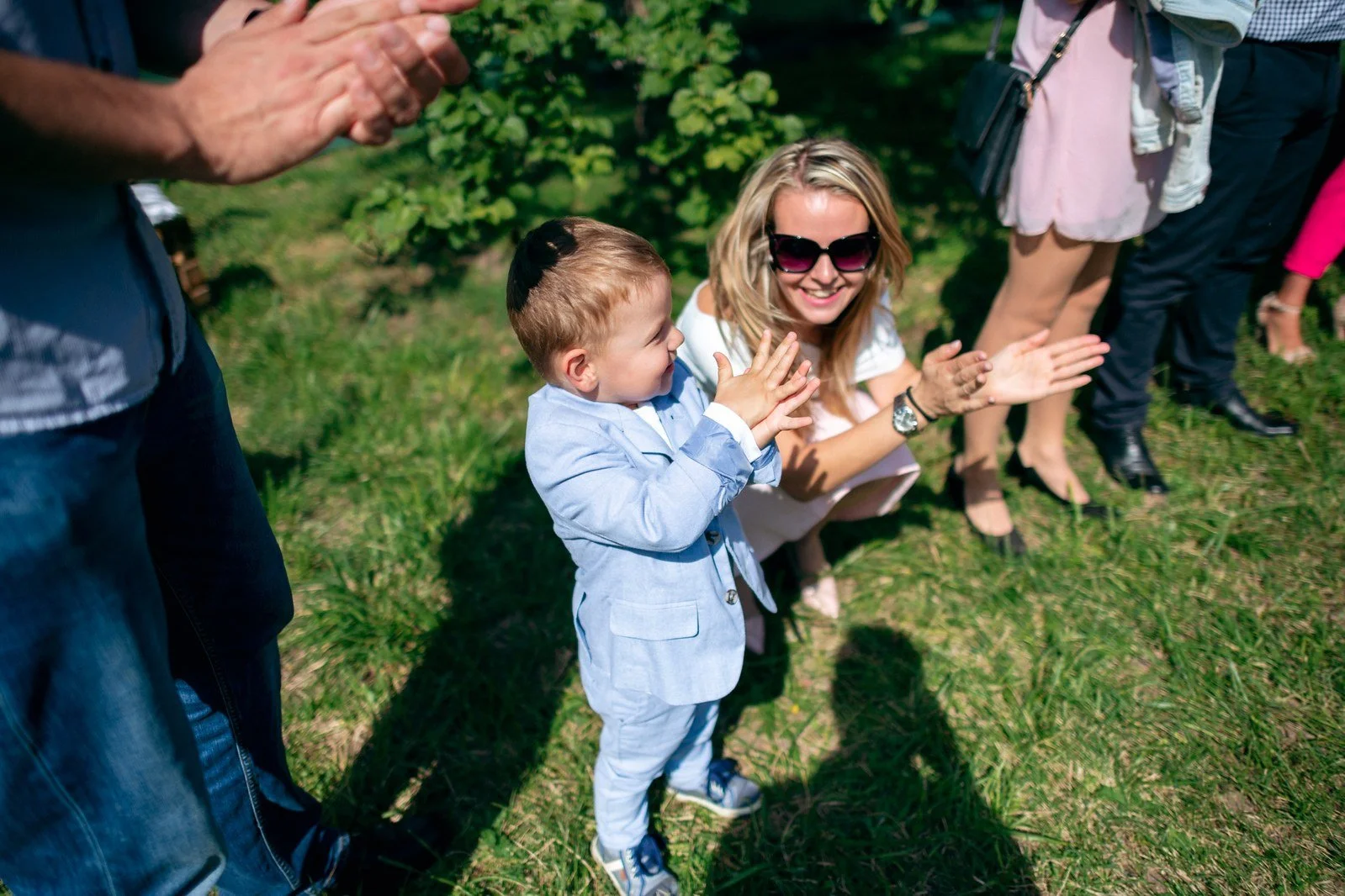Chlapec ve světle modrém obleku tleská vedle klečící usměvavé blondýnky se slunečními brýlemi.