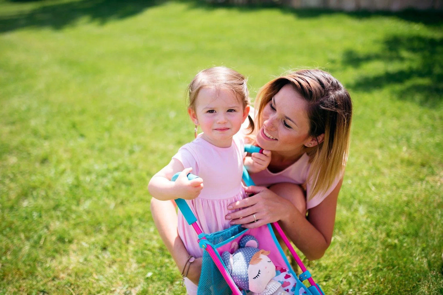 Malá dívka drží madla barevného kočárku pro panenky a žena v růžových šatech se k ní sklání na trávníku.