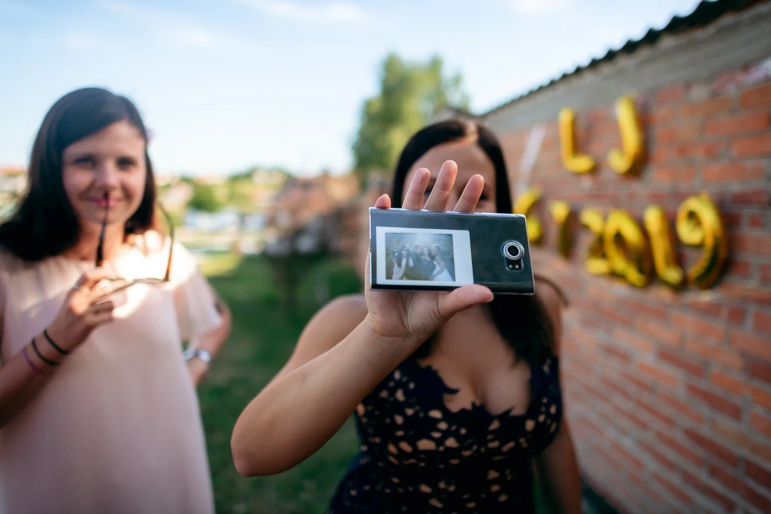 Žena v popředí míří instantním fotoaparátem k objektivu, druhá drží drobný předmět u cihlové zdi s výzdobou.