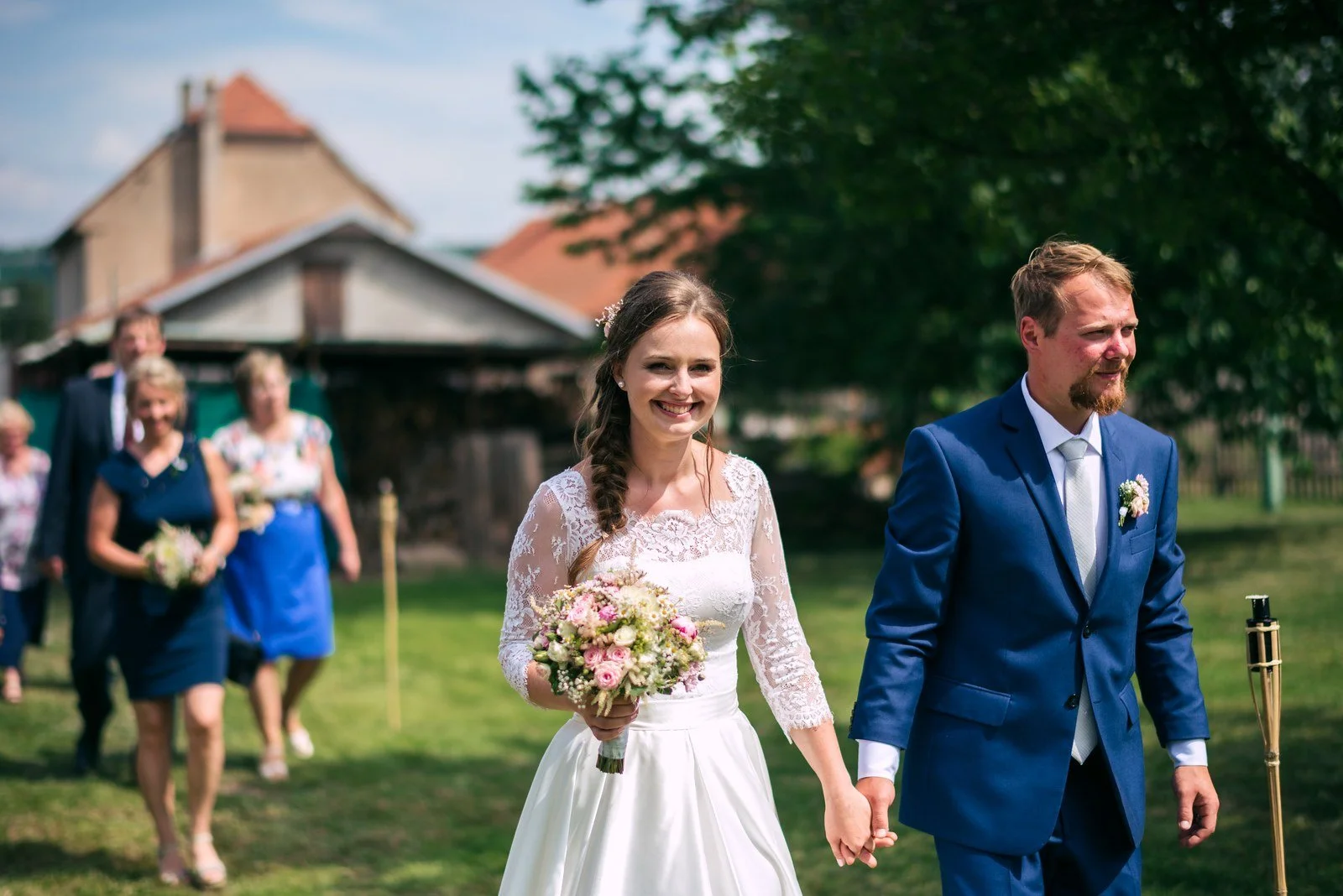 Žena s kyticí a muž v modrém obleku kráčejí po zahradě.