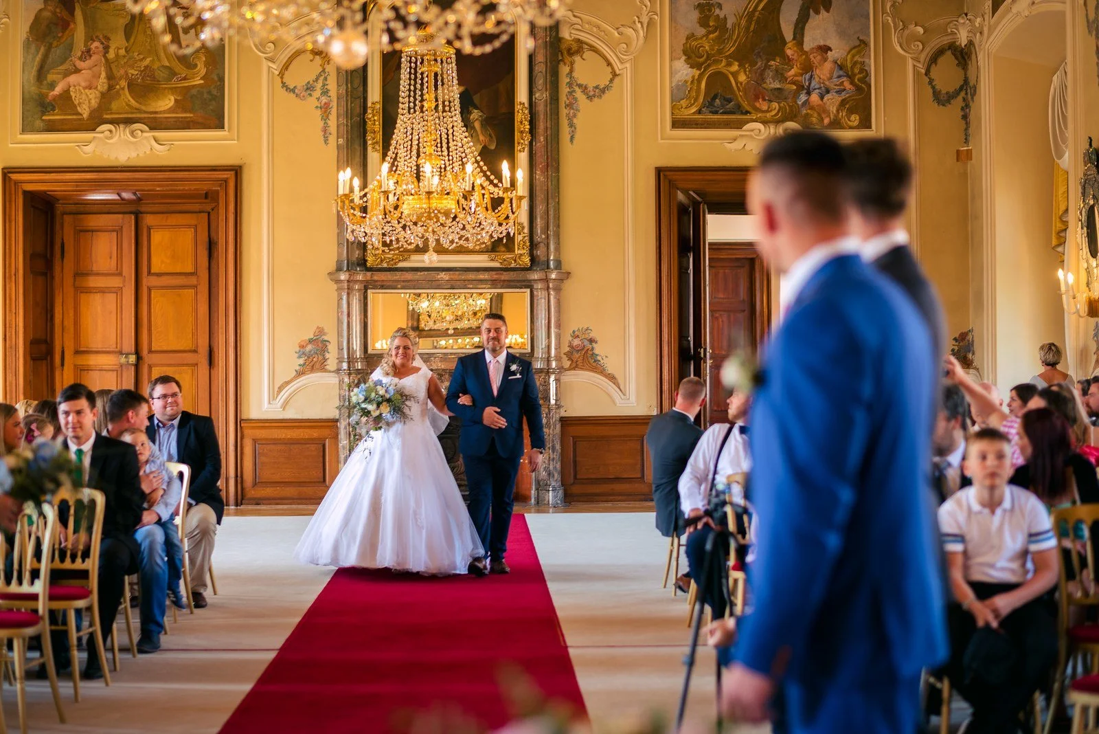 Pohled přes ženichovo rameno na příchod nevěsty po červeném koberci.