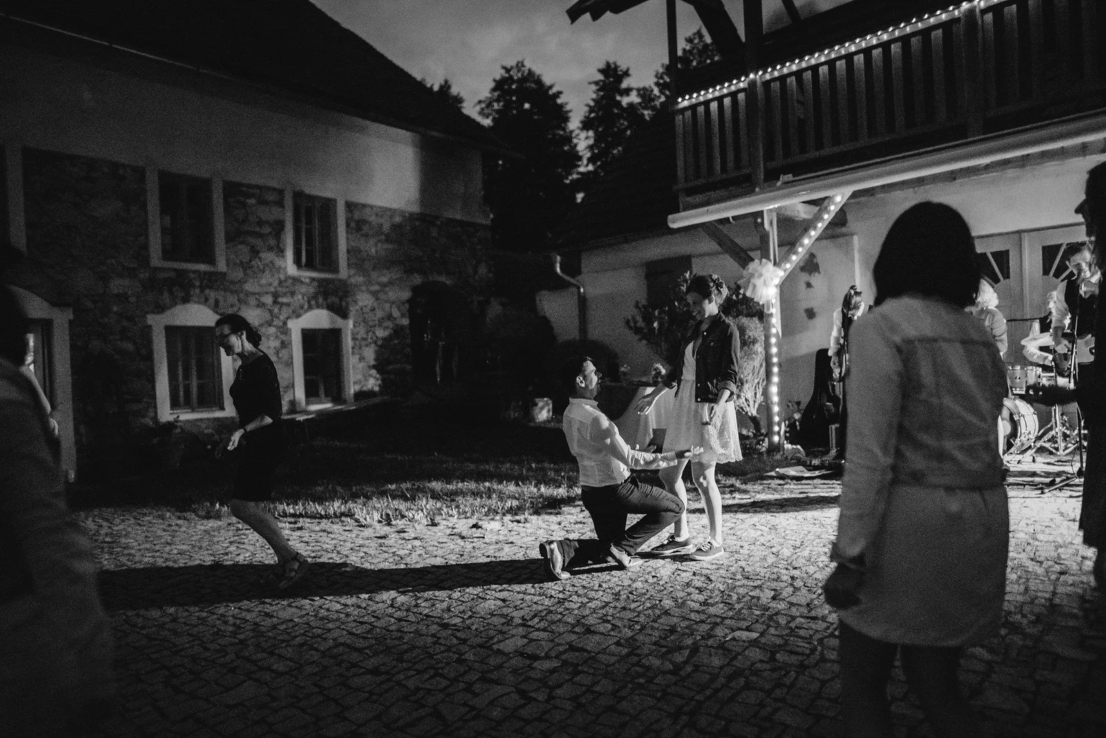 Černobílá noční fotografie zachycuje pohybující se postavy mezi historickými budovami.