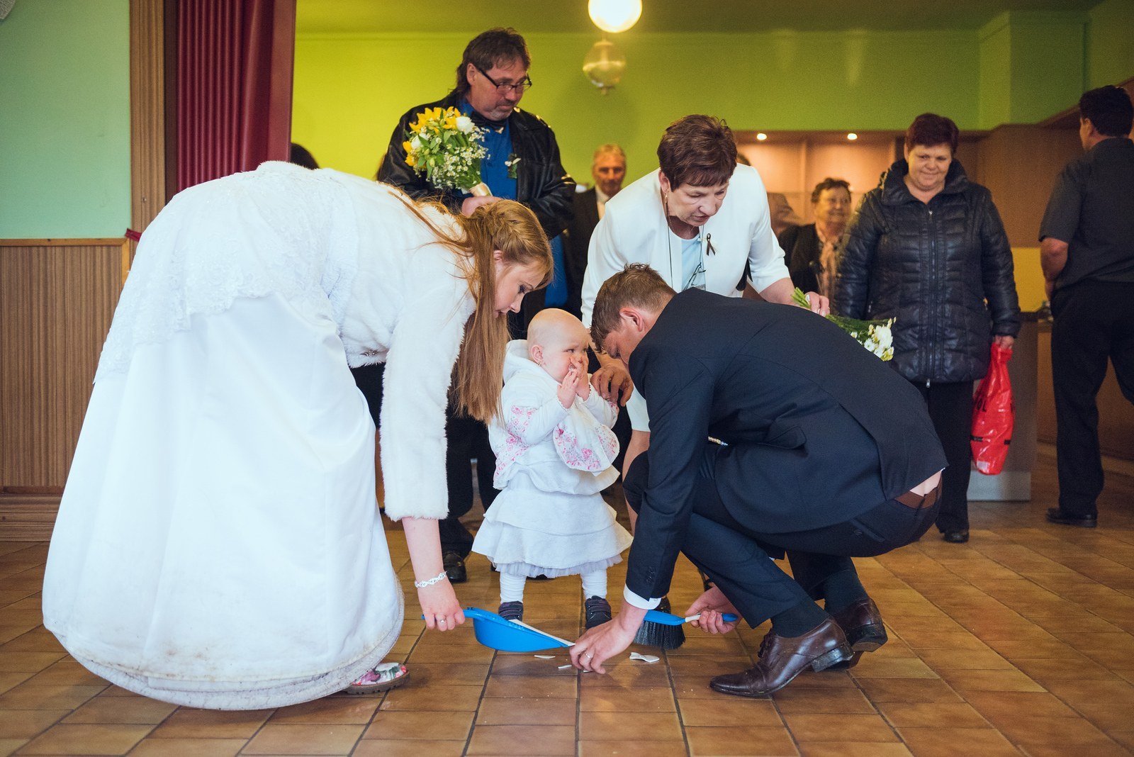 Nevěsta a ženich zametají střepy z podlahy vedle malé dívky v bílých šatech.