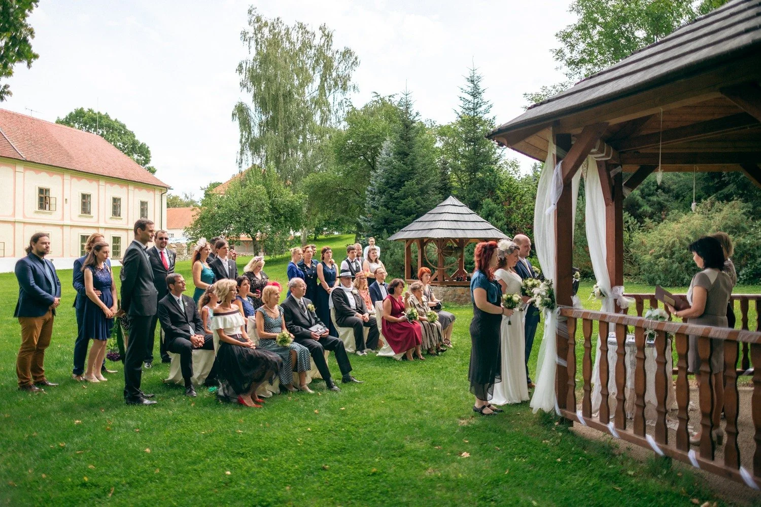 Hosté sedí na zahradě před altánem připraveným pro obřad.