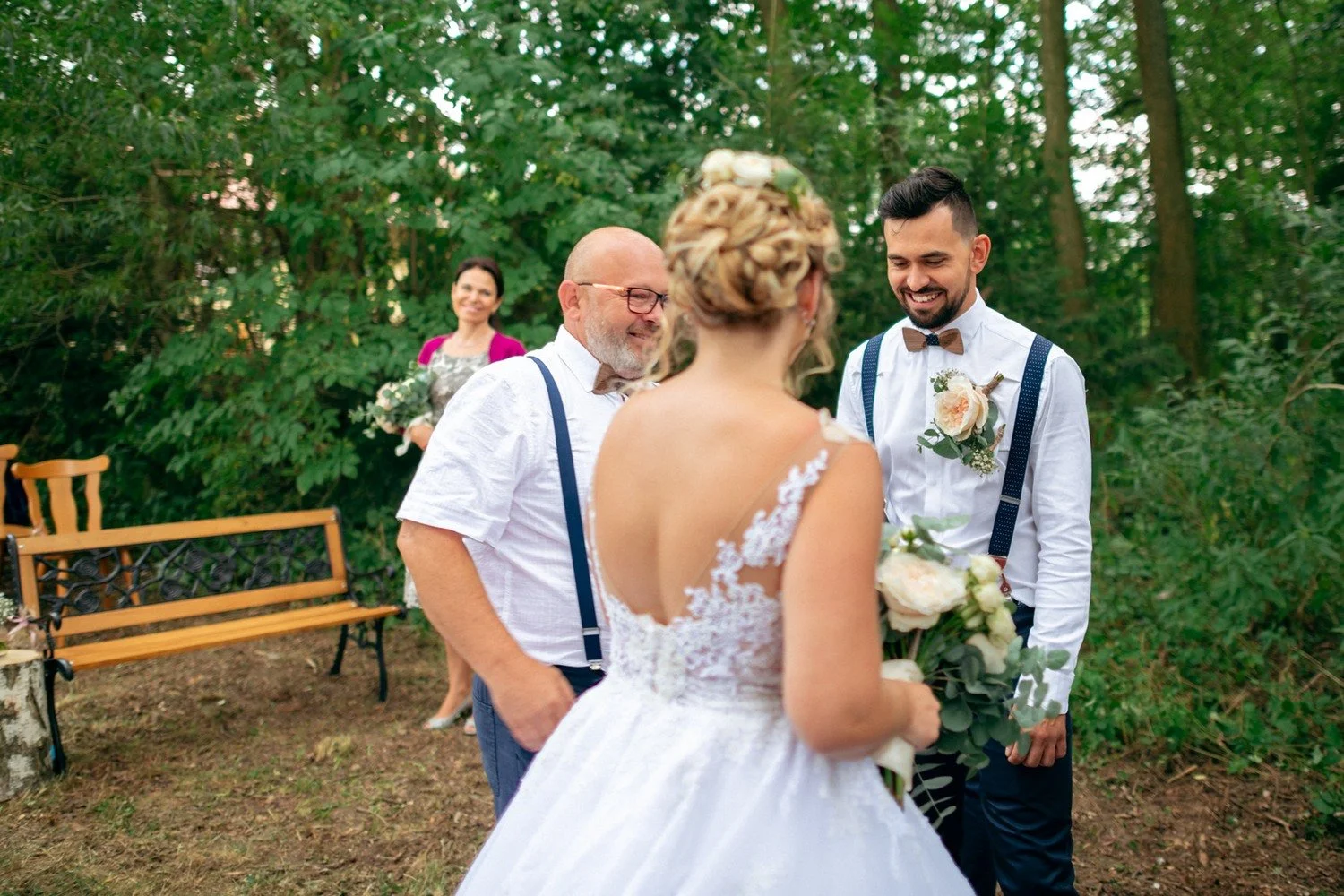 Nevěsta stojí zády k fotoaparátu před ženichem a mužem s bílými vlasy.