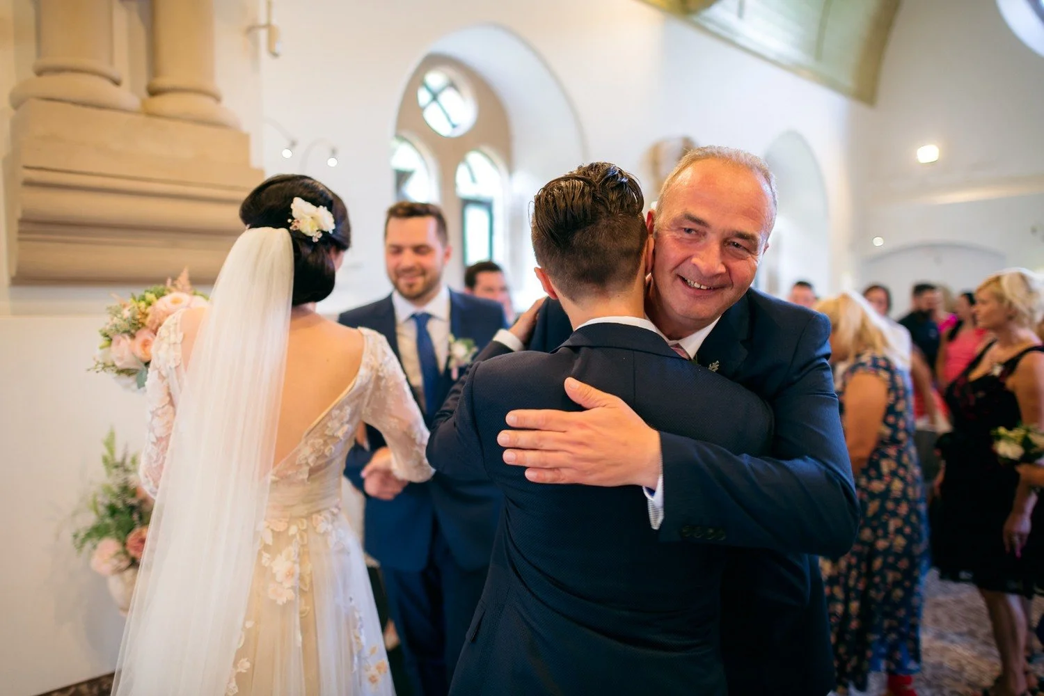 Usmívající se muž v tmavém obleku objímá muže otočeného zády, vedle nich stojí nevěsta s dlouhým závojem.