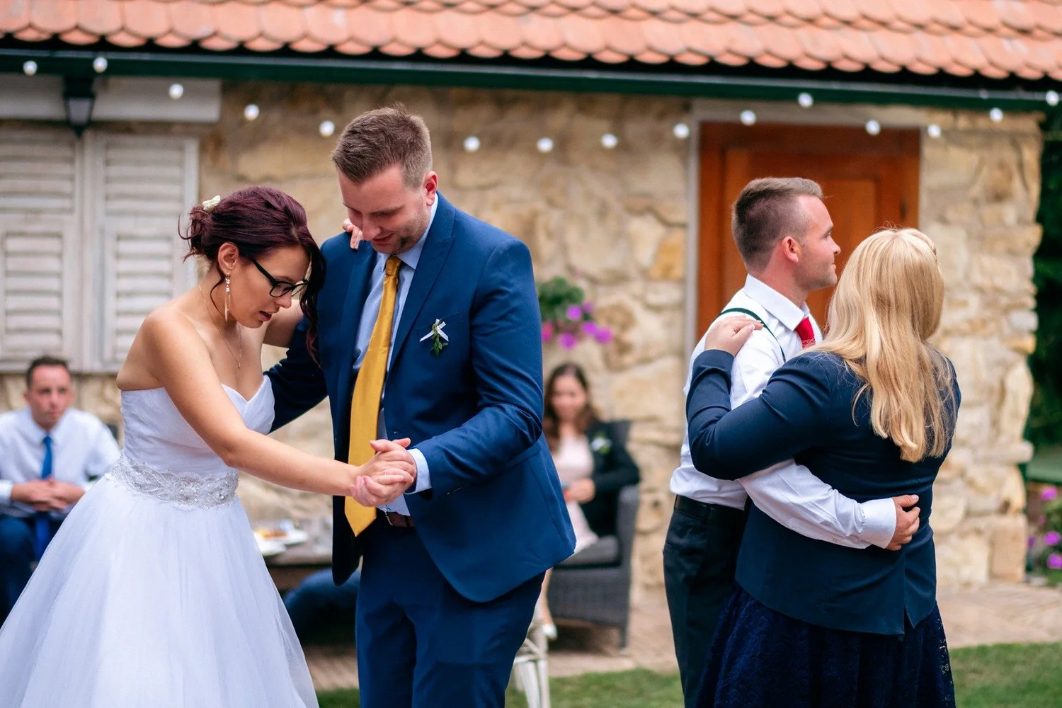 Žena v bílých šatech tančí s mužem v modrém obleku před kamennou budovou.