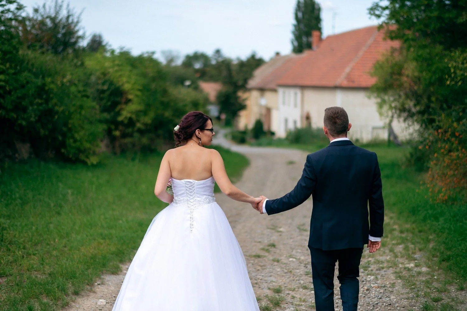 Pár zachycený zezadu jde ruku v ruce po cestě mezi trávou a keři.