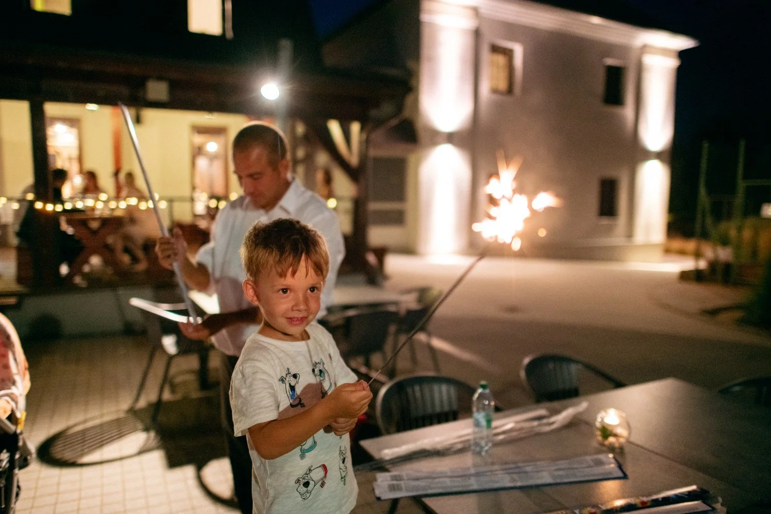 Dítě s prskavkou v popředí a rozsvícený penzion za ním uvádějí závěrečný motiv s místním kontextem.