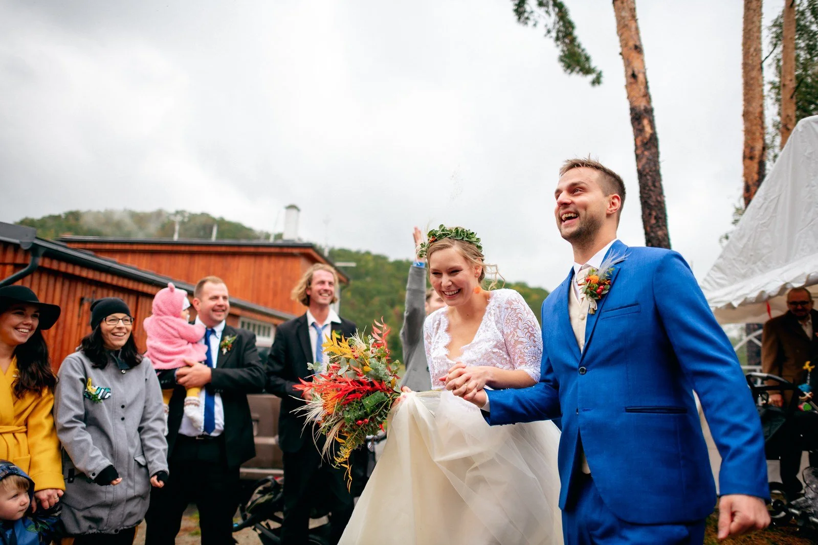 Žena s kyticí a muž v modrém obleku vycházejí ven kolem skupiny přihlížejících.