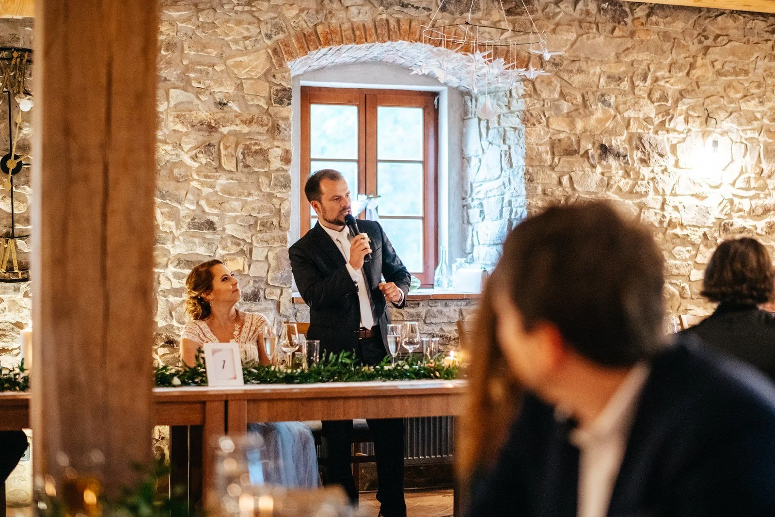 Muž v tmavém obleku stojí s mikrofonem před hosty v kamenném sále.