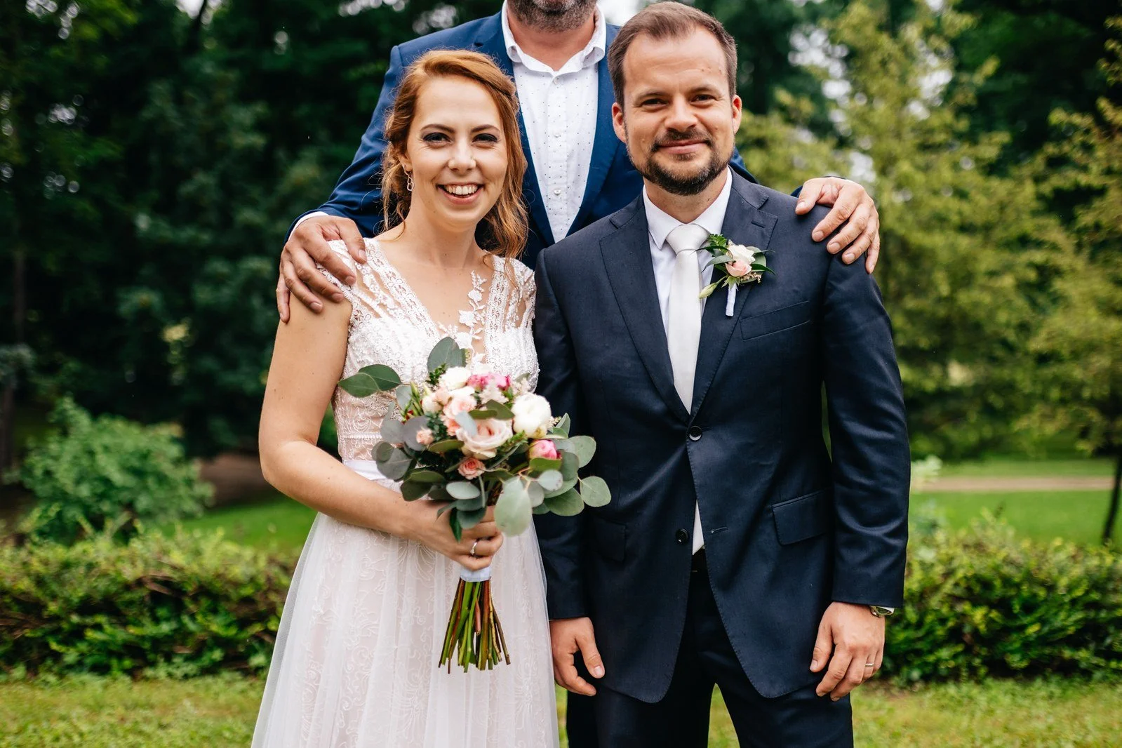 Dvojice stojí vedle sebe na trávníku před hustou zelení, žena drží kytici a muž má tmavý oblek.