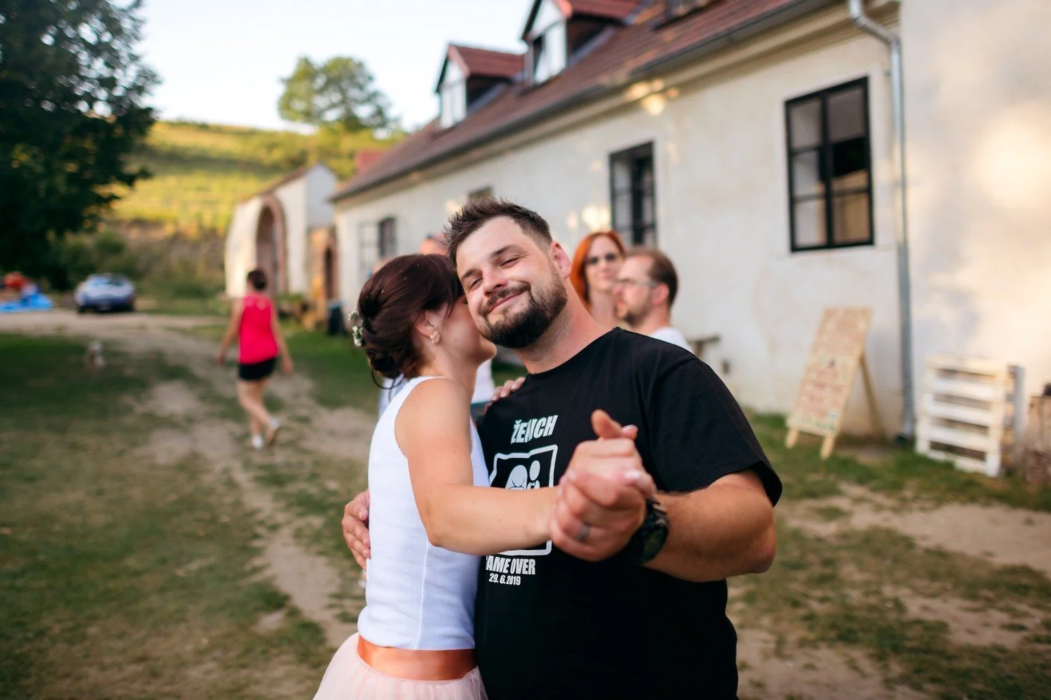 Usměvavý muž v černém tričku tančí v objetí se ženou před bílou budovou.