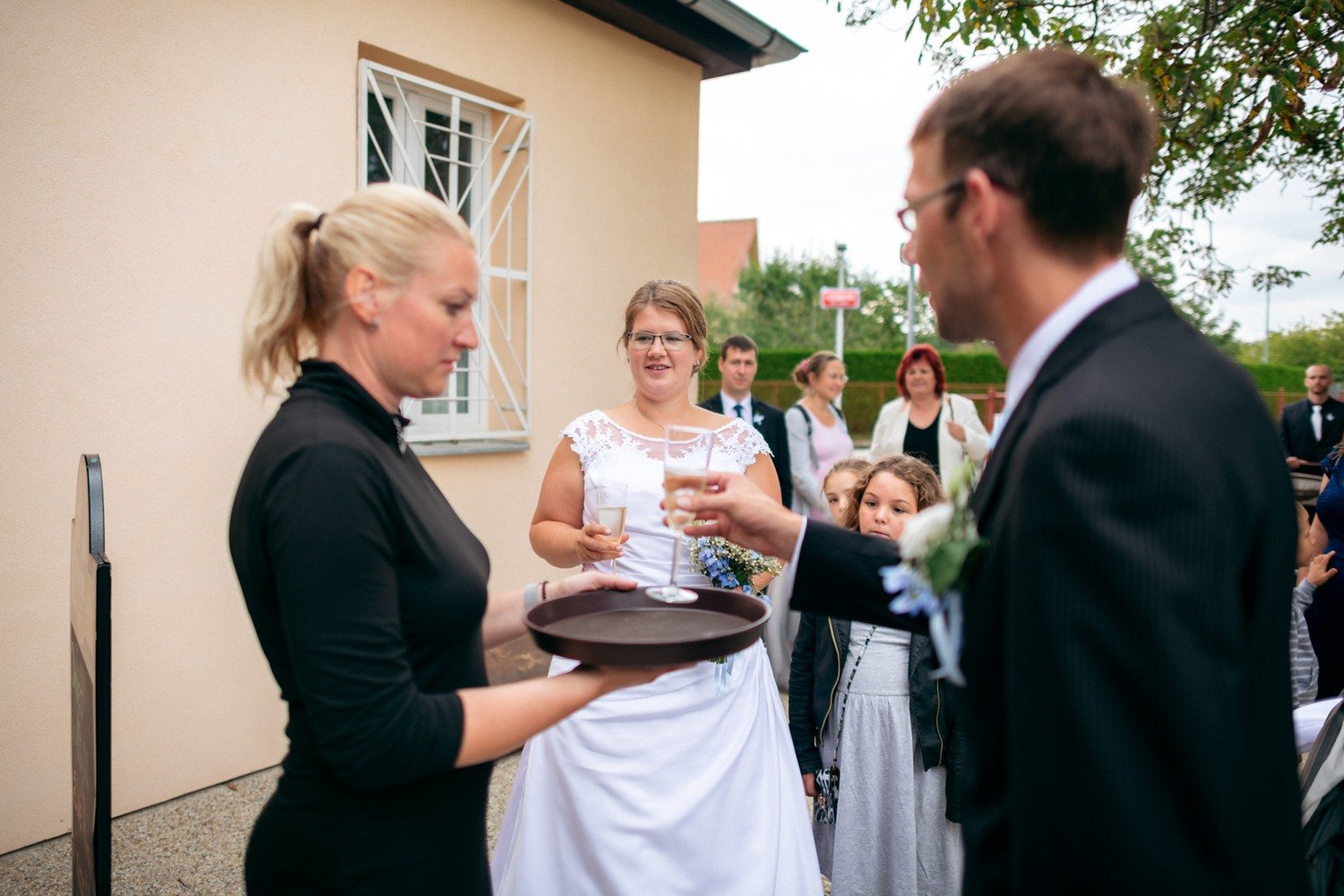 Žena v černém podává sklenku muži v obleku před skupinou hostů.