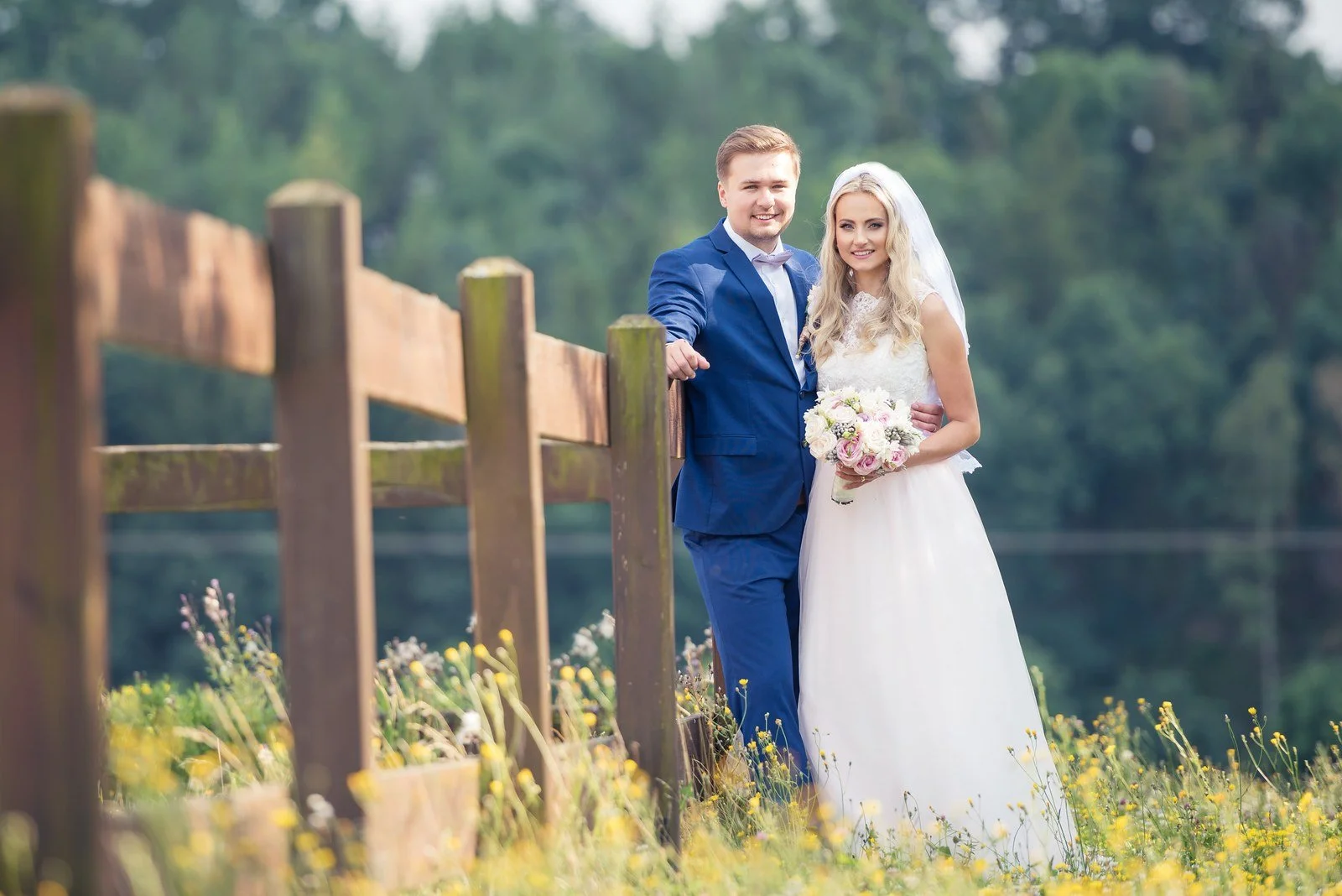 Nevěsta a ženich stojí v trávě vedle dřevěného ohradníku.