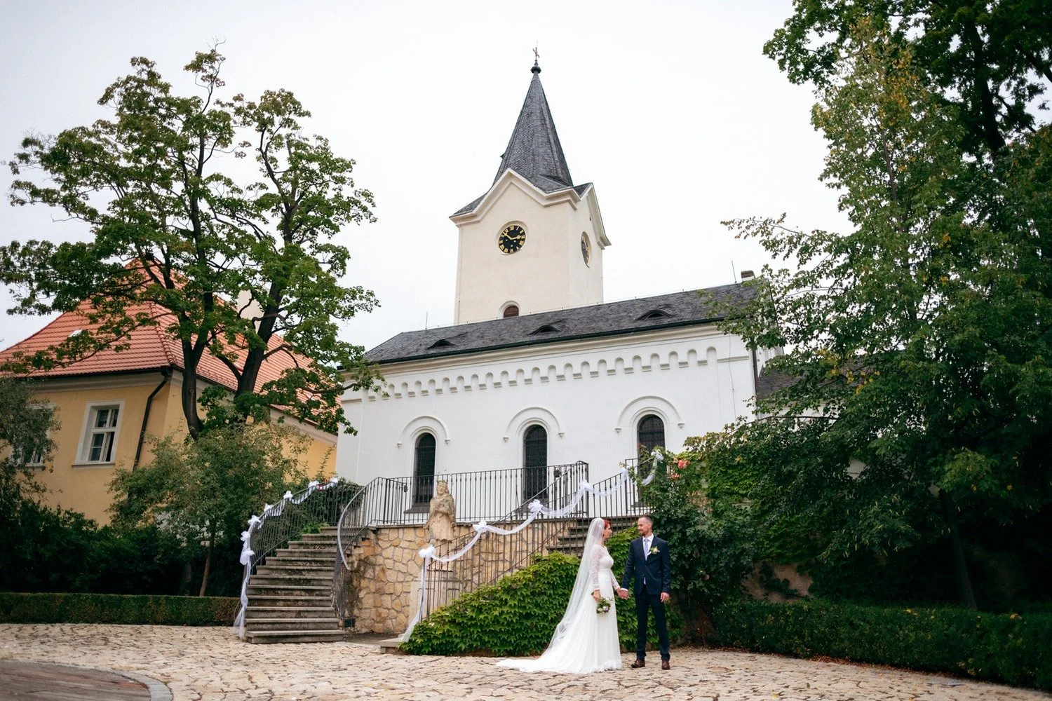 Novomanželé stojí před bílou kaplí obklopenou stromy.