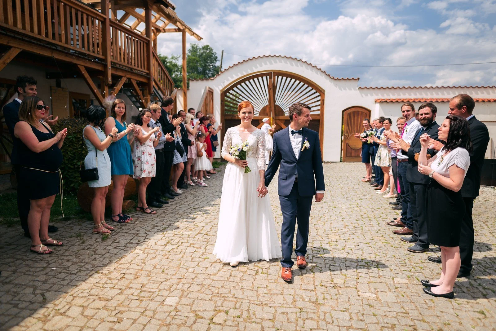 Dvojice prochází dřevěným obloukem mezi lidmi, kteří stojí podél dlážděné cesty.